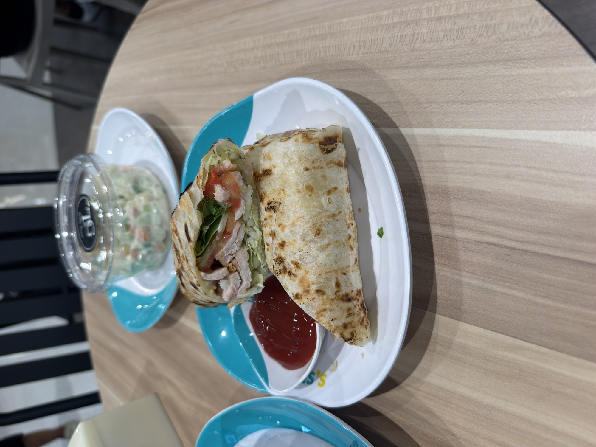 In the city of Ban Rang Rak, Thailand, a close-up, high-angle shot captures a meal on a wooden table. The primary subject is a wrap cut in half, revealing fillings of sliced meat, lettuce, and tomato, placed on a white plate with a teal rim. Beside it is a small dish of ketchup. In the background, a clear plastic container holds coleslaw, also on a white plate with a teal rim. The table surface is made of light-colored wood planks with a visible grain. The lighting is ambient, suggesting an indoor setting. No people are clearly visible in the frame.
