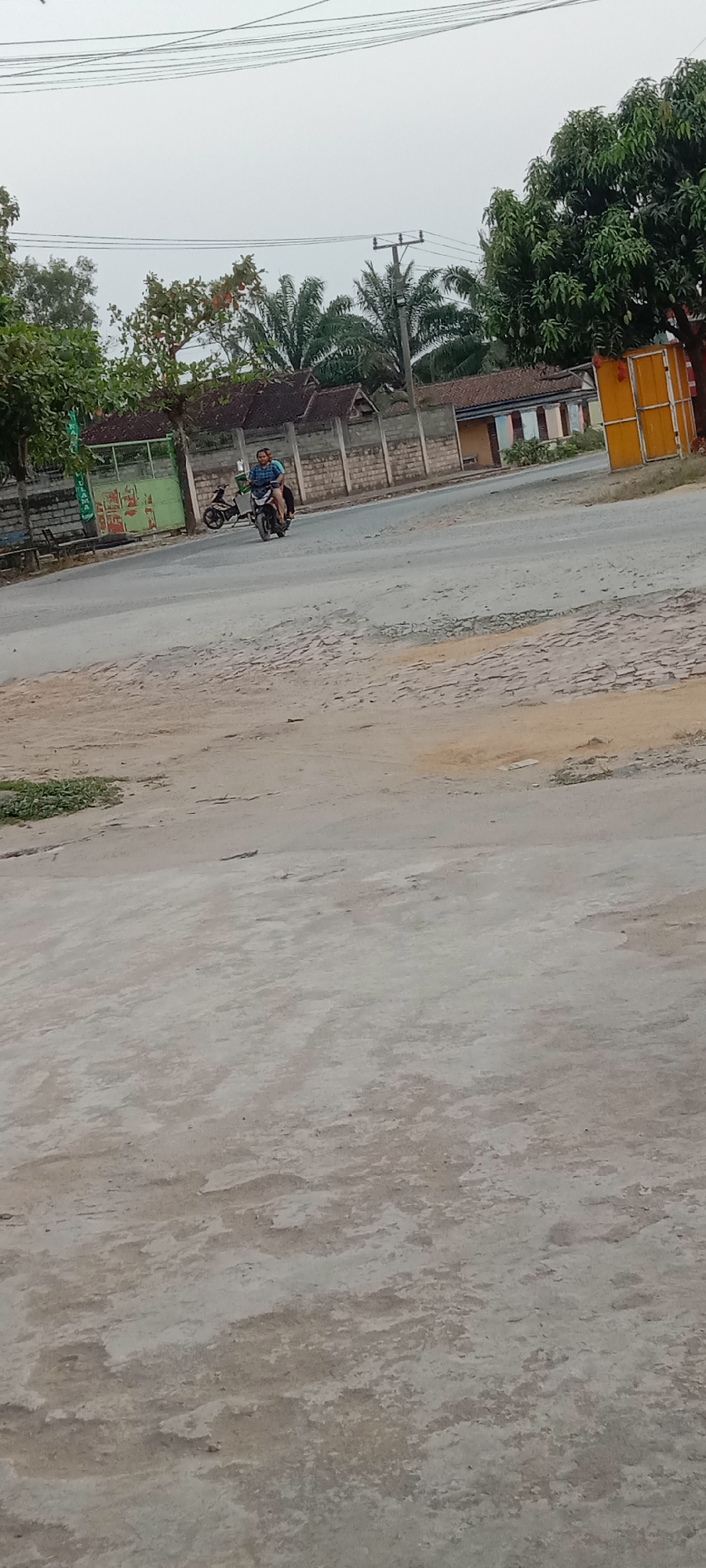A person is riding a motorcycle on a paved road in a semi-urban area. The person is wearing a blue and white checkered shirt. A second, parked motorcycle is visible to the left of the person on the road. In the background, there are buildings with tiled roofs and a fence. Trees, including palm trees and a leafy tree, are present. A utility pole with overhead wires stands near the buildings. The sky is overcast. The foreground consists of a dirt and concrete surface. The scene appears to be in Utama Jaya, Mataram, Indonesia, based on the context provided.