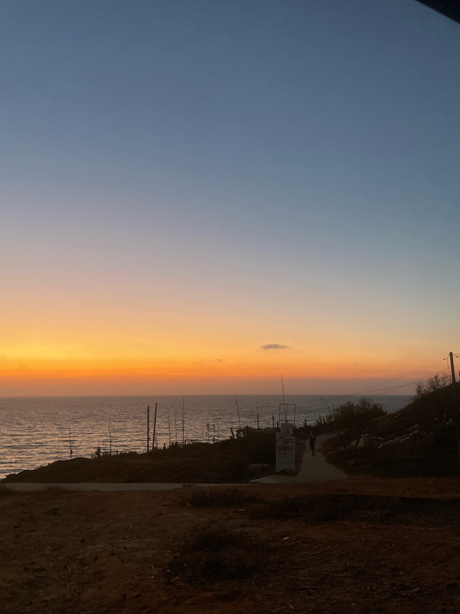 The image depicts a twilight scene over the Mediterranean Sea, likely taken from a coastal path in Herzliya, Israel. The sky displays a gradient of colors, transitioning from a deep blue at the top to warm orange and yellow hues near the horizon, indicative of sunset or sunrise. The sea reflects these colors, with gentle waves lapping at the shore.
In the foreground, a rough, earthy terrain with sparse vegetation leads to a paved pathway. Along the pathway, a lone figure is walking towards a signpost. To the right of the path, a small hill is covered with rocks and scrubby bushes, topped by a utility pole with electrical wires.
On the left side of the image, overlooking the sea, are several tall, slender poles, possibly remnants of old structures or fishing equipment. Some sparse greenery and darker shrubbery are visible along the coastline. A sign board stands near the pathway, adding a touch of human presence to the natural landscape. The overall atmosphere is serene and tranquil, capturing a moment of natural beauty by the coast.