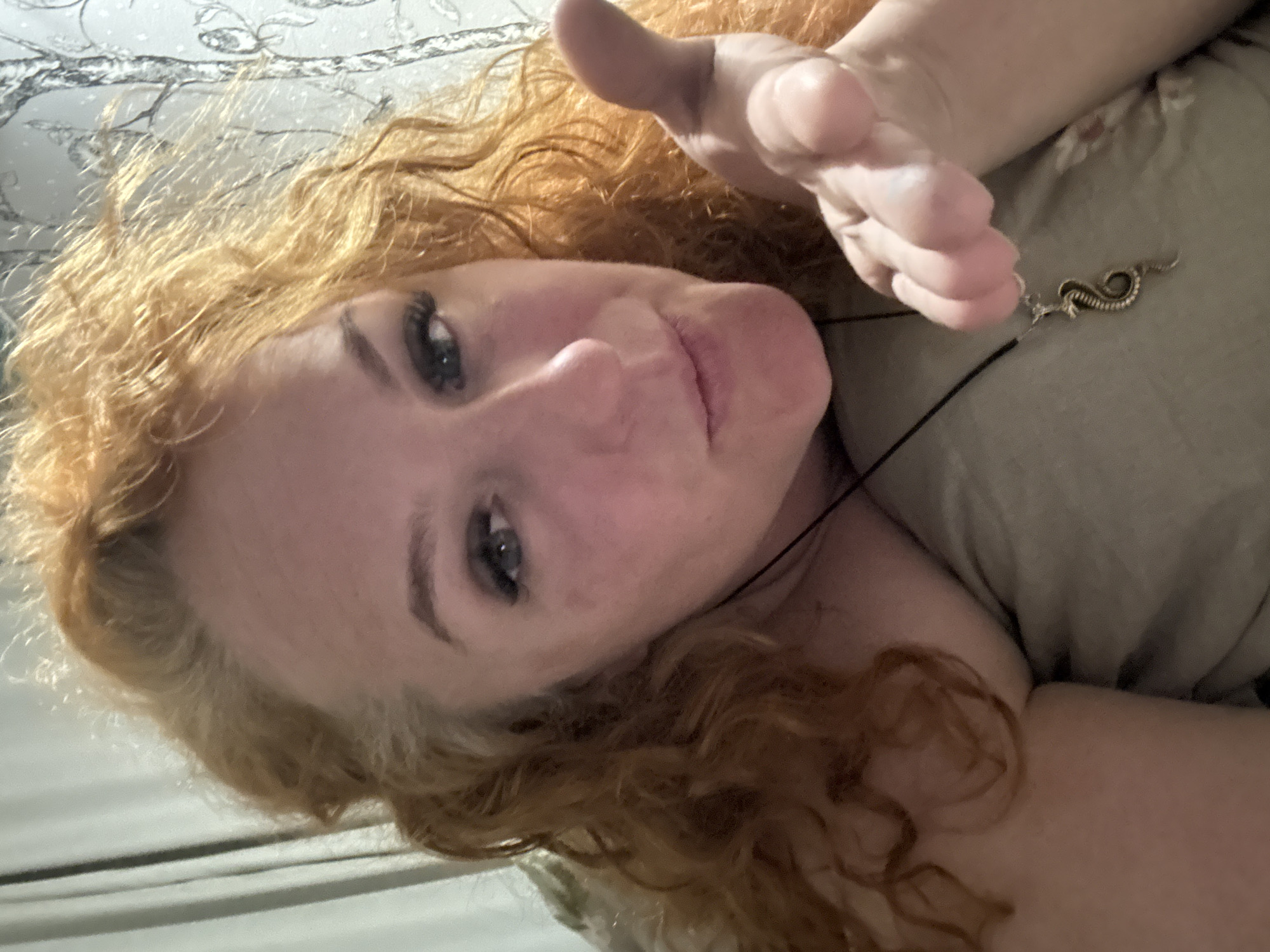 A selfie depicts a woman with light complexion, red curly hair, and blue eyes. She is wearing a black cord necklace with a serpentine pendant. Her hand is extended towards the camera with fingers slightly curled. The background includes a surface with a light, abstract pattern and the edge of what appears to be a white surface. The image does not contain any information related to the location in the city of Chaikovsky, Russia.