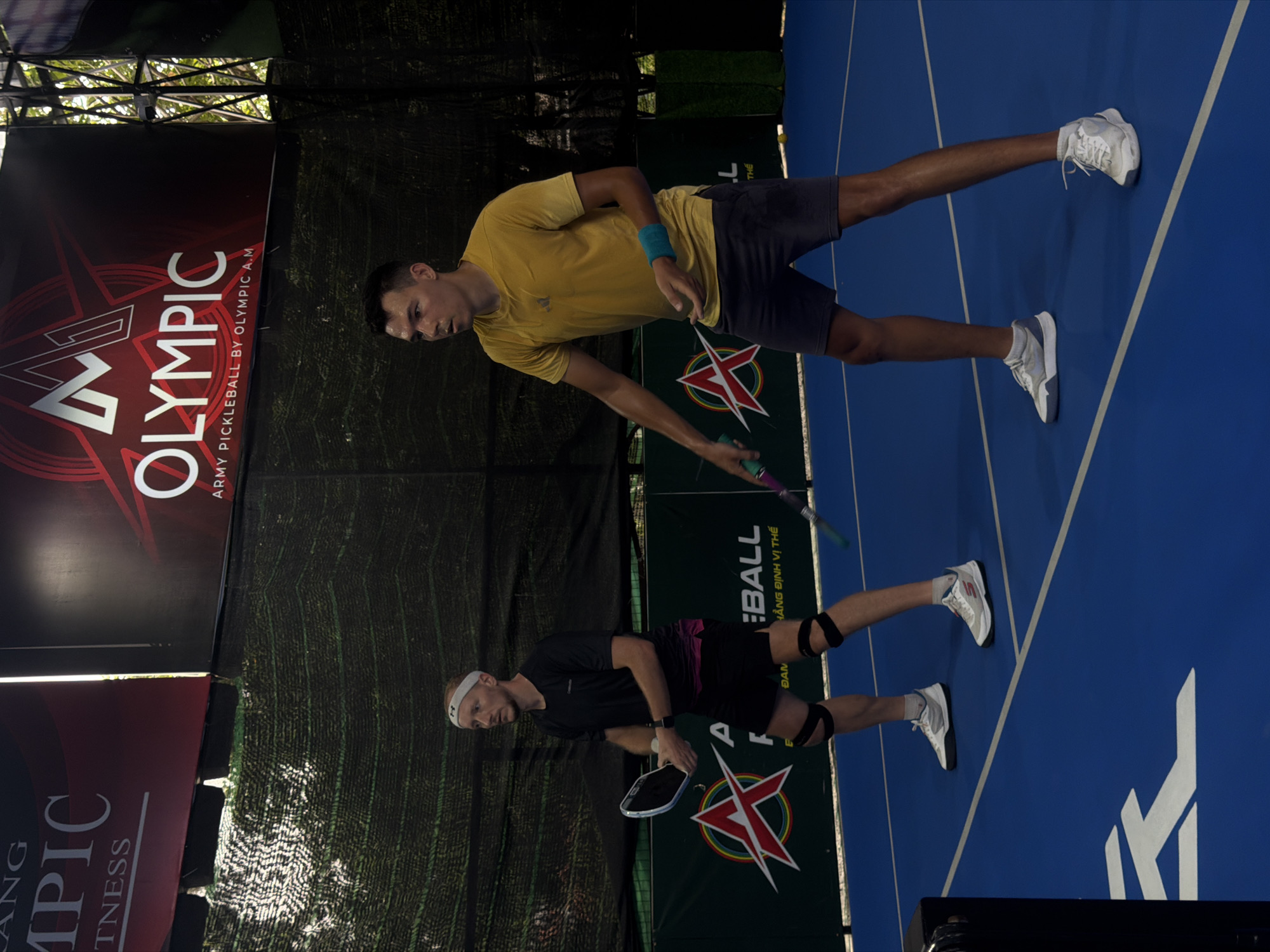 Here is a description of the image:
The image shows two men playing pickleball on a blue court with white lines. One man is wearing a yellow shirt and dark shorts, and the other is wearing a dark shirt and dark shorts with a white headband. They are both holding pickleball paddles and appear to be in the middle of a game.
Behind the men, there are banners with the words "OLYMPIC" and "ARMY PICKLEBALL BY OLYMPIC A.M." on them. The banners also have a logo with a star and the words "PICKLEBALL HẰNG ĐỊNH VỊ THẾ" in Vietnamese.
The location is in Bac Nha Trang Ward, Vietnam, as evidenced by the Vietnamese text on the banners and other visual cues.