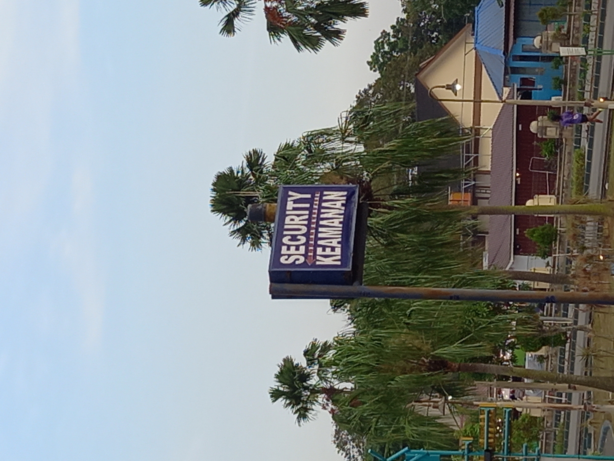 The image shows a blue sign with white text that reads "SECURITY KEAMANAN" and an arrow pointing to the left. The sign is mounted on a dark pole and is surrounded by palm trees and other vegetation. In the background, there are several buildings, including one with a light-colored roof and another with a blue roof. A person is seen running on a path in the middle ground. The sky is clear and light blue. The scene appears to be outdoors, possibly in a park or public area. The presence of the sign and the general atmosphere suggest a security checkpoint or an area requiring surveillance. The architecture and vegetation are consistent with a location in Samarinda, Indonesia, a city known for its tropical climate and palm trees.