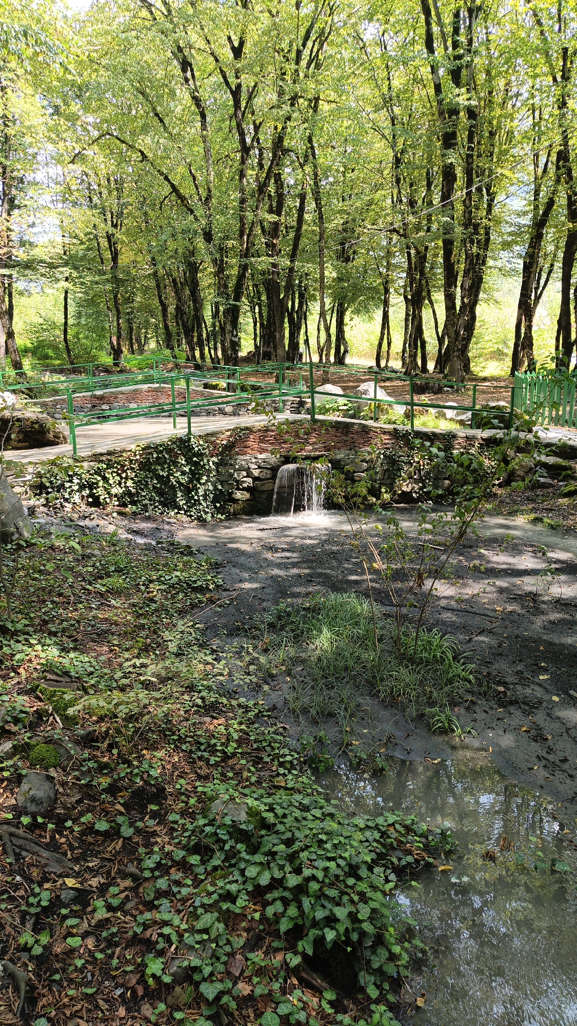 The image depicts a picturesque scene in Gabala, Azerbaijan, featuring a small waterfall flowing from a brick-lined structure into a muddy stream. A charming brick bridge with green railings spans the stream, surrounded by lush greenery and tall trees. The ground is covered with fallen leaves and ivy, adding to the natural beauty of the setting. The overall ambiance is serene and tranquil, evoking a sense of peace and quiet.