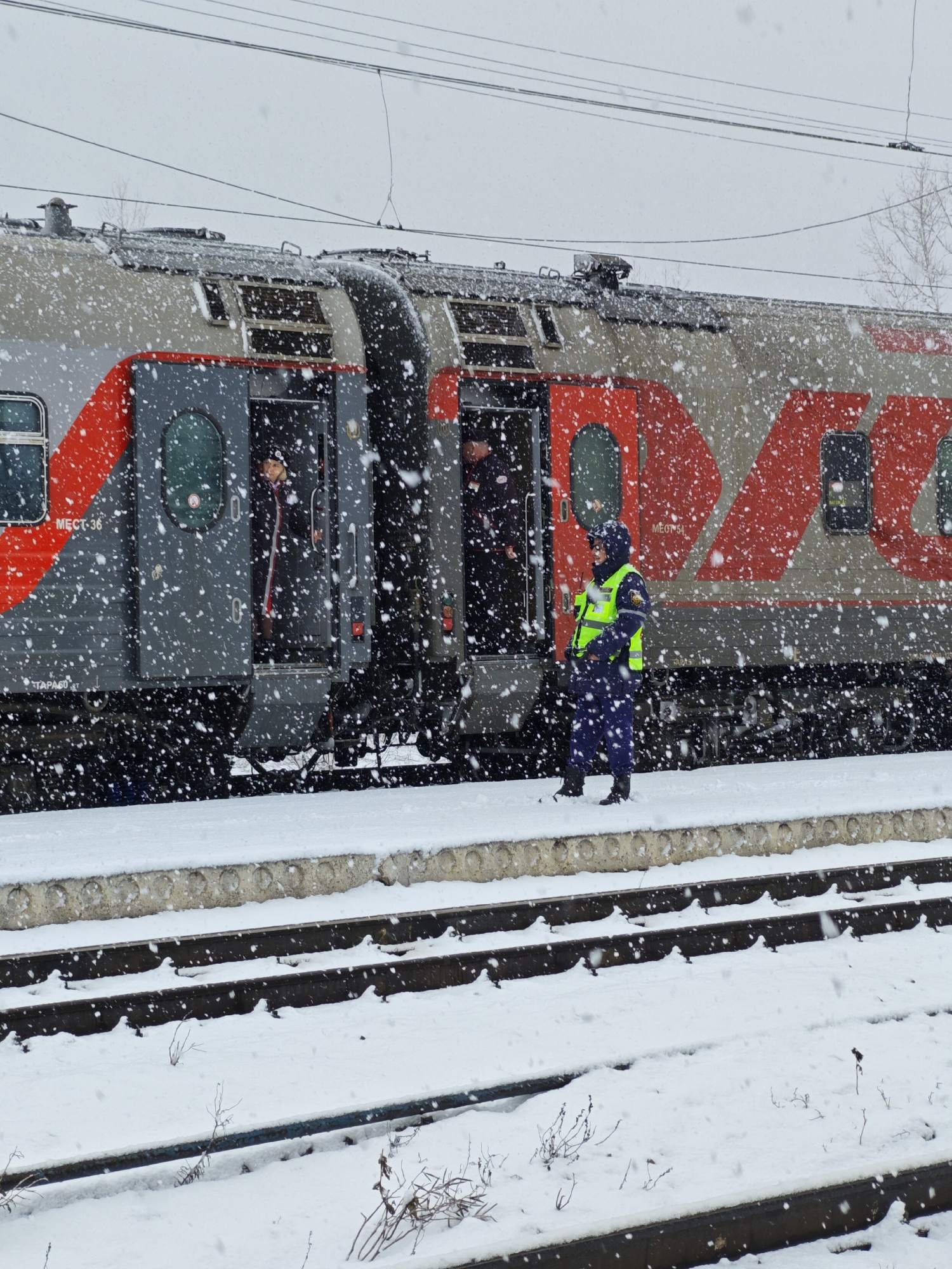 The image captures a snowy scene at what appears to be a train station in Kambarka, Russia. A red and gray Russian Railways (RZD) train is stopped at a platform. Two figures can be seen in the train doorways. A rail worker wearing a bright green safety vest and dark blue uniform stands on the platform next to the train. Heavy snowfall is visible, obscuring some details. The train tracks in the foreground are partially covered in snow. The environment is wintery and overcast.