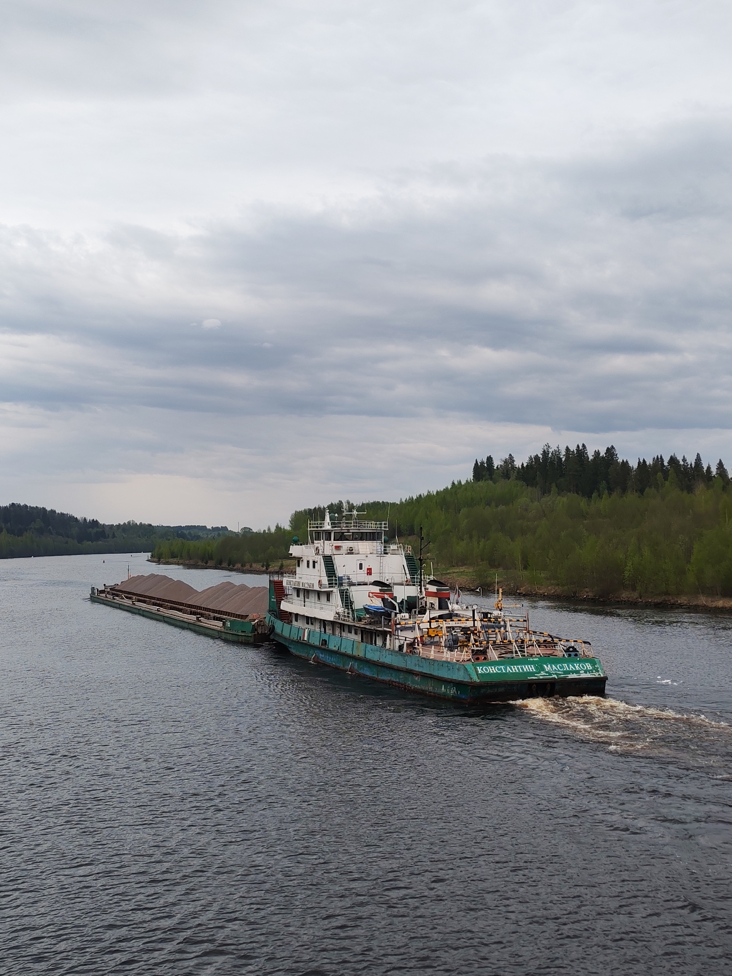 A wide shot depicts a tugboat and barge on a body of water. The tugboat, painted in teal and white, is labeled "Константин Маслаков" in Russian. A barge laden with what appears to be brown gravel or ore is attached to the tugboat's side. Behind the water, a forested shoreline is visible under a cloudy sky. The scene is likely located near Alekseevskoe, Russia.