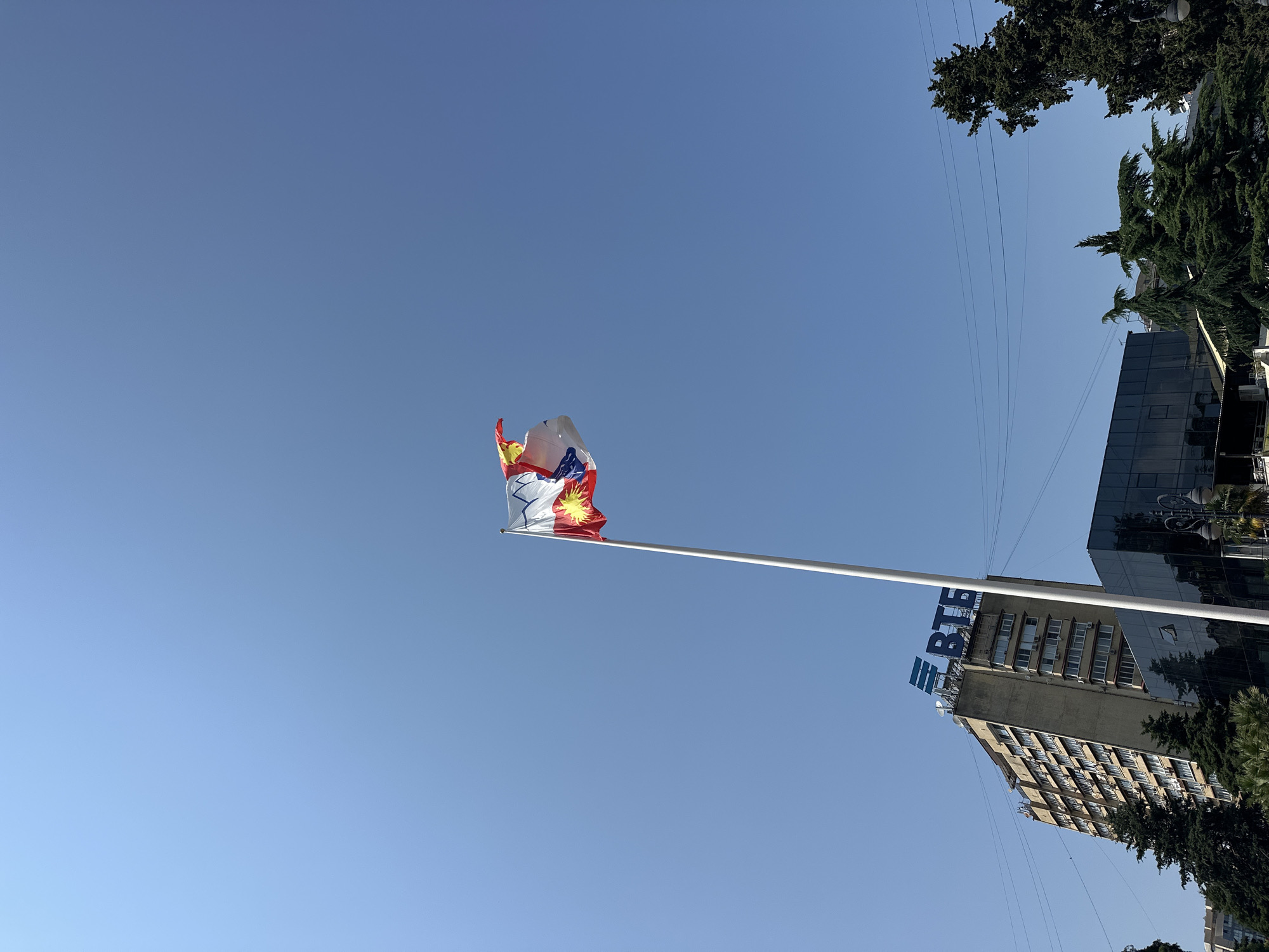 The image depicts a bright, sunny day in Sochi, Russia, with a clear blue sky dominating the upper portion. A tall white flagpole stands prominently in the center, with a colorful flag waving in the breeze. The flag features a white background with blue and red elements, and a depiction of a sun with rays.
Below and to the left of the flagpole, a multi-story building with a beige facade and many windows is visible. The building has a sign on its upper portion that reads "ВТБ" in large blue letters, indicating it is a branch of VTB Bank. Adjacent to this building, to its right, is a modern building with a dark, reflective glass facade.
Several tall, dark green evergreen trees are scattered throughout the scene, adding a natural element to the urban landscape. Overhead power lines are also visible, crisscrossing the sky. The overall impression is a clear, summery day in a city setting, with a focus on the flag and the surrounding architecture.