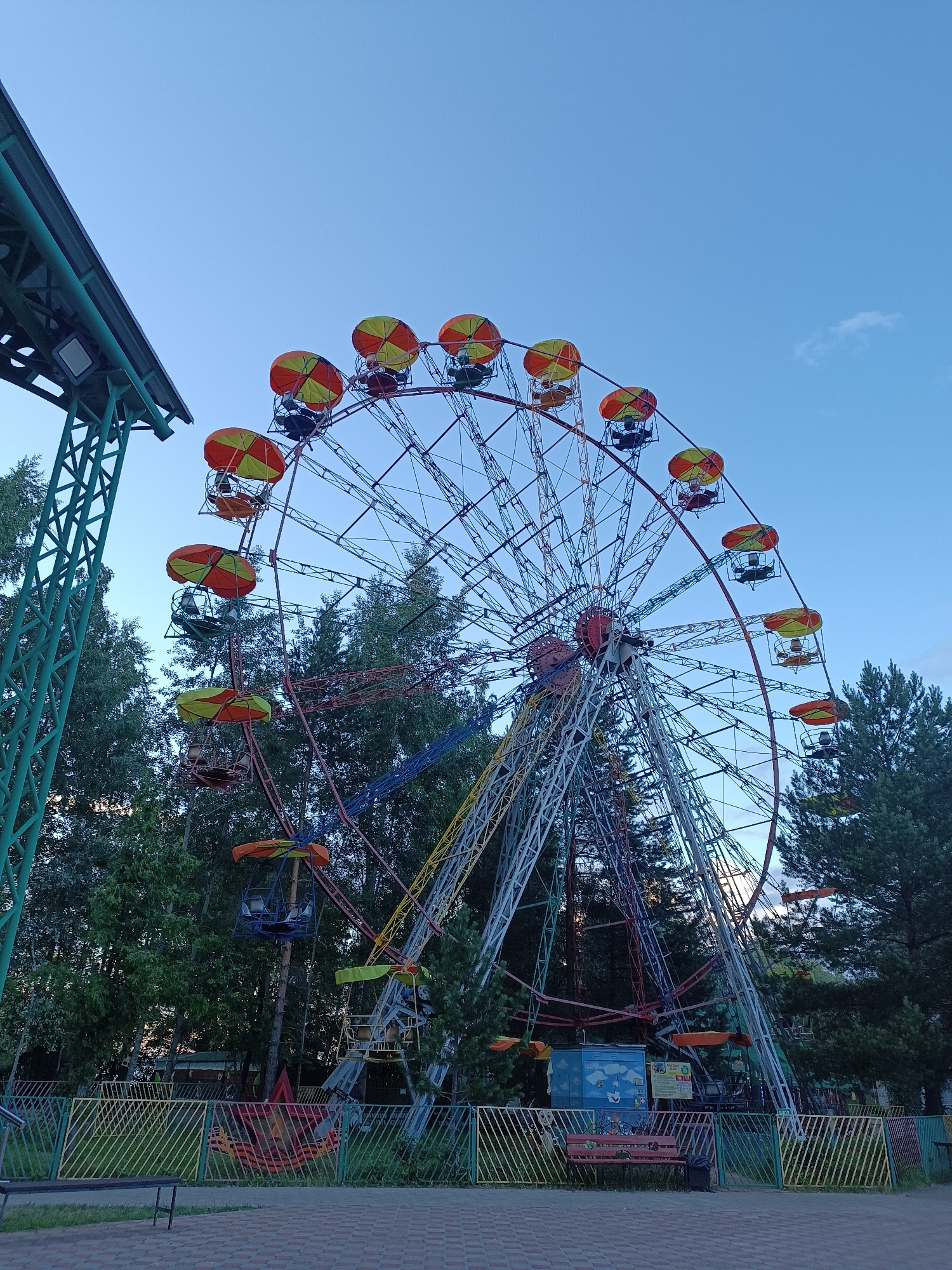 An amusement park Ferris wheel with orange and yellow canopy-covered cars stands against a clear blue sky. The Ferris wheel is a complex metal structure with multiple support beams and radiating spokes. Several cars are visible, each with passengers. In the foreground, there is a green metal structure with a canopy, possibly part of the park's infrastructure, and a low-lying fence with yellow and green metal pickets. Beyond the fence, trees form a dense backdrop. The ground is paved with interlocking bricks. The scene appears to be in an outdoor amusement park. The location is identified as Chaikovsky, Russia.