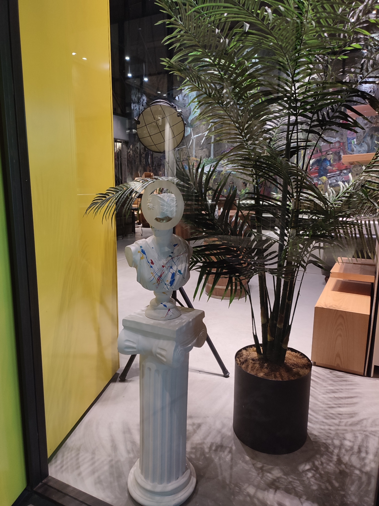 A white bust, splattered with red, blue, and yellow paint, sits on a pedestal. The bust features a halo-like ring above its head. To the right of the bust is a large potted plant with lush green fronds. The scene is framed by a bright yellow wall to the left and a dark, reflective surface, likely glass, to the left and top. In the background, through the glass, are hints of an indoor environment with furniture and decorative elements. The floor is a light gray, with shadows cast by the plant and other unseen objects. This appears to be an interior scene, possibly a shop or exhibition space, located in Sarıyer, Turkey.