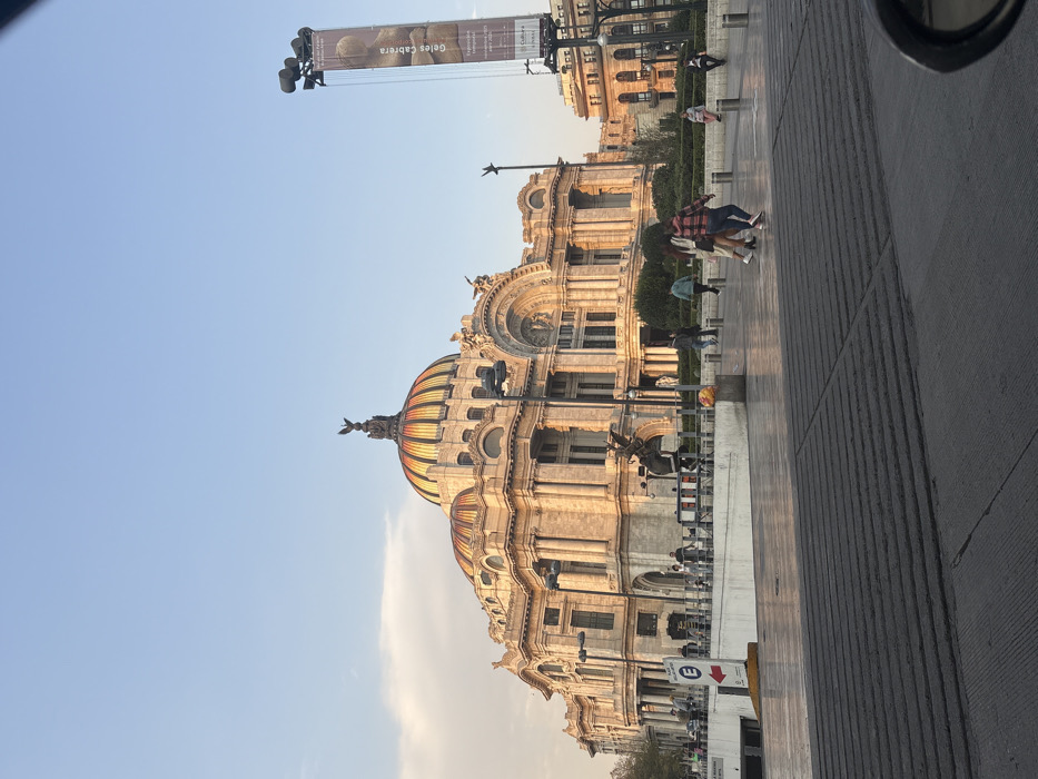 The Palace of Fine Arts in Mexico City, featuring a distinctive dome with vibrant colors, occupies the central visual space. The building's ornate facade is bathed in golden light, suggesting either sunrise or sunset. A tall advertising banner stands adjacent to the palace. People walk along the adjacent sidewalk. A street sign and the edge of a vehicle are in the foreground. The sky is blue, adding contrast to the scene.