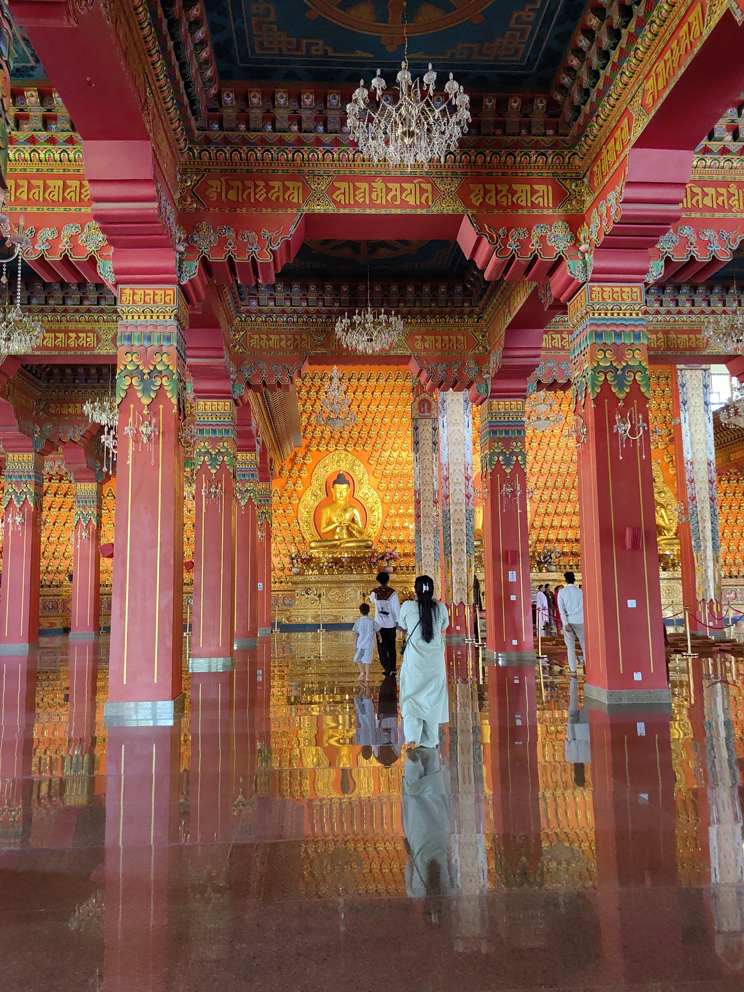 The image depicts the interior of a Buddhist temple, likely located in Parsa, Nepal, given the context. The hall is grand and ornate, with vibrant red pillars adorned with gold trim and colorful, intricate carvings. The ceiling features decorative elements and chandeliers. In the background, a large golden Buddha statue sits prominently on an altar, surrounded by a wall covered in numerous smaller golden Buddha figures. The floor is highly polished and reflective, mirroring the pillars and the gilded walls, creating a sense of depth and grandeur. Several individuals are present in the hall, some walking and others standing, observing the central Buddha statue. The overall atmosphere is one of reverence and architectural splendor.