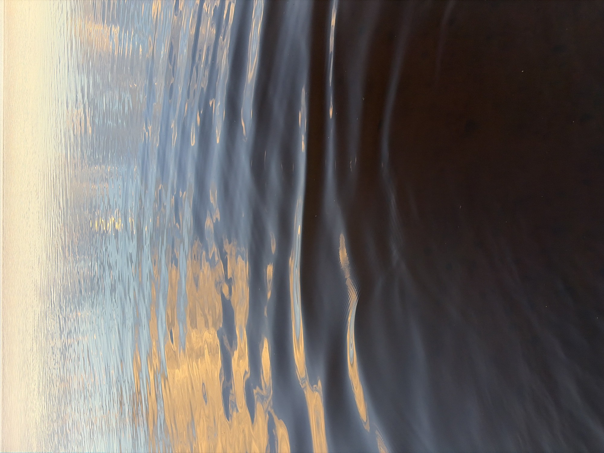 A close-up, high-angle shot shows the surface of dark, rippling water. The water is reflecting the sky, with hues of pale yellow, orange, and light blue visible in shimmering patches on the surface. The ripples create a textured pattern across the water. The bottom portion of the image is a deep, dark brown, with some lighter brown reflections. There are a few small white specks scattered across the surface, possibly debris or insects. The overall impression is one of a calm, natural body of water at dawn or dusk.