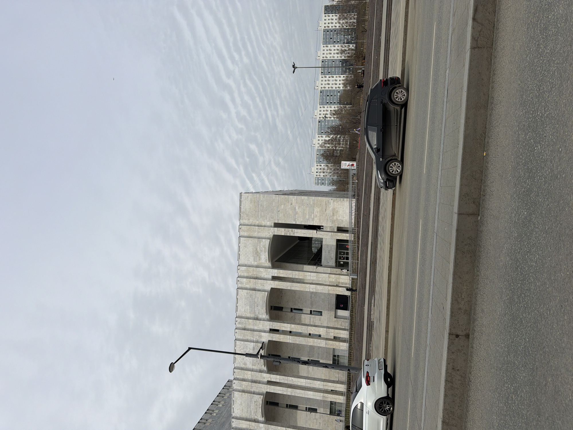 The image depicts a street scene in Perm, Russia, with a multi-lane road flanked by buildings. The building on the left appears to be a large, modern structure constructed of light-colored stone or concrete, featuring a series of arched facades. A streetlamp stands in front of the building. On the right side of the road, high-rise apartment buildings are visible in the background. Two cars are present: a white car closer to the camera and a dark car further down the road. The sky is overcast with a textured cloud pattern. No people are readily identifiable in the image.