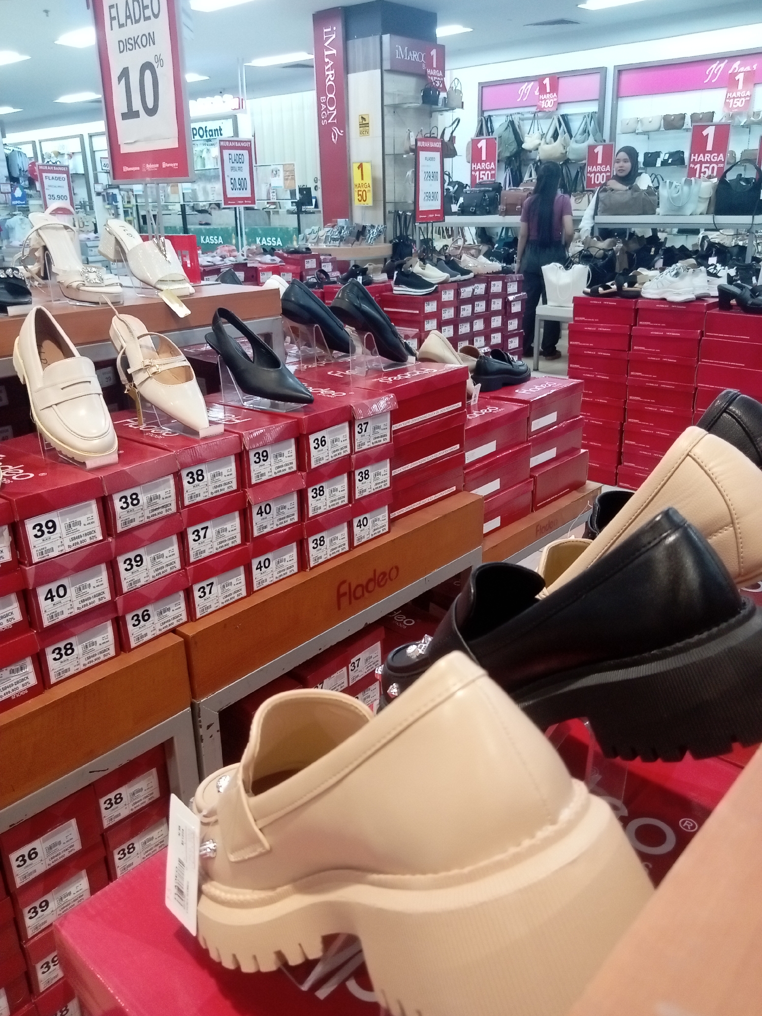 This image captures a retail environment, likely a shoe store, with a focus on displaying various footwear and handbags. The scene is organized with tiered shelving units and red cardboard boxes stacked high, suggesting a large inventory. Several pairs of shoes, including loafers, heels, and chunky-soled platforms, are prominently displayed on clear acrylic stands. Some shoes are presented with price tags and discount information, such as "FLADEO DISKON 10%" and "HARGA 50.900". Handbags are visible on shelves in the background, with some bearing signage like "IMAROCON BAGS". The lighting appears to be typical of indoor retail spaces, with overhead fluorescent lights. The presence of Indonesian currency and language on the price tags, along with the signage, indicates the location is in Indonesia. Given the retail context and common store chains, this scene is plausibly from a department store or a dedicated shoe store in Batam, Indonesia, a city known for its shopping destinations. There are no people clearly visible in the foreground, but a woman is partially visible in the mid-ground.