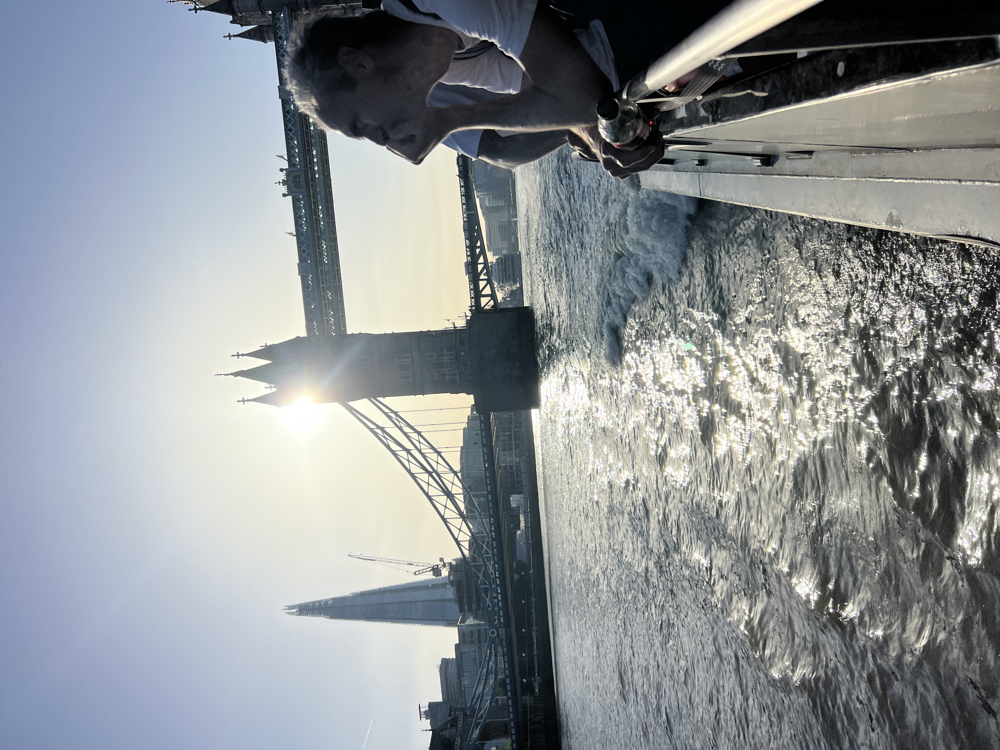 A person is leaning over a railing, looking down at the water, with the Tower Bridge and The Shard in the background. The scene takes place on a sunny day in London, United Kingdom, likely along the River Thames.