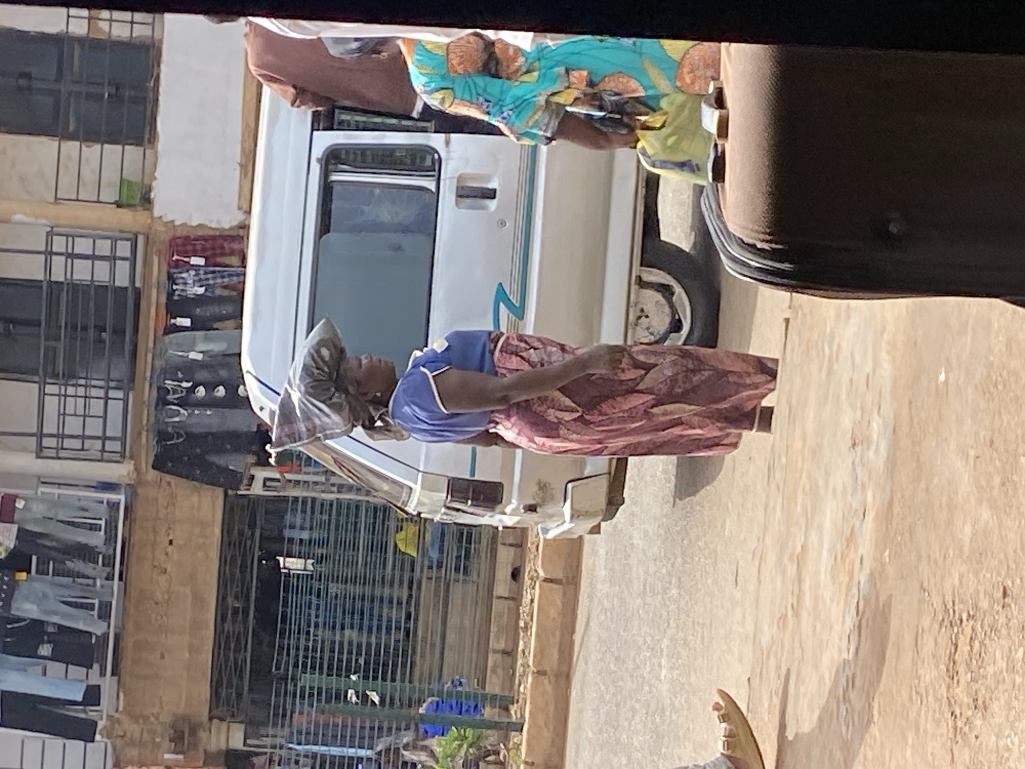 In Bukuru, Nigeria, a woman walks on a dirt road next to a silver minivan. She carries a large bundle wrapped in fabric on her head. She wears a blue shirt and a long, patterned skirt. Another woman, partially visible, stands to the right of the minivan wearing a colorful, patterned dress. In the background, a building with multiple windows and balconies is visible, with clothing hanging from racks. The scene depicts a typical street view in a Nigerian city, with people going about their daily activities.
