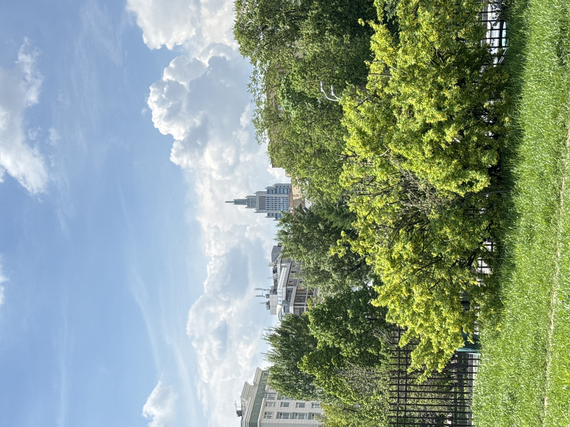 The image shows a view of the sky with puffy white clouds and a tall building in the background, partially obscured by trees. In the foreground, there is greenery, likely a park or garden, with a fence visible. The architecture of the building suggests it might be one of the Stalinist skyscrapers. The location is most likely Moscow, Russia, given the architectural style of the tall building. There are no people in the shot. The environment appears to be a sunny day in a city park.