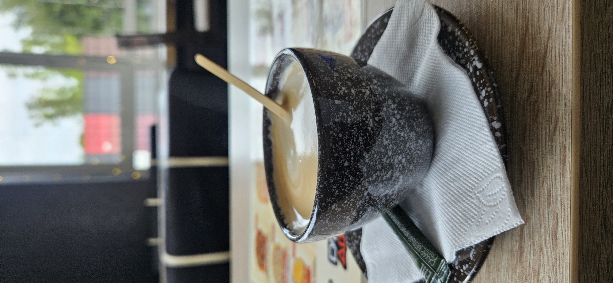 A close-up, eye-level shot shows a dark speckled ceramic bowl filled with a frothy beverage, likely coffee or latte, with a wooden spoon partially submerged in it. The bowl rests on a matching ceramic saucer, which is covered by a white paper napkin. A single-serving packet of sugar, partially obscured by the napkin and spoon, lies on the saucer. The setting appears to be a cafe or restaurant with a blurred background featuring large windows that reveal greenery and distant buildings, suggesting an urban environment. The table surface is made of light-colored wood. The image is sharpest on the bowl and its contents, with a shallow depth of field blurring the surroundings. There are no people visible. This scene does not contain any specific visual indicators to place it in Vitsebsk, Belarus; it depicts a generic cafe setting.