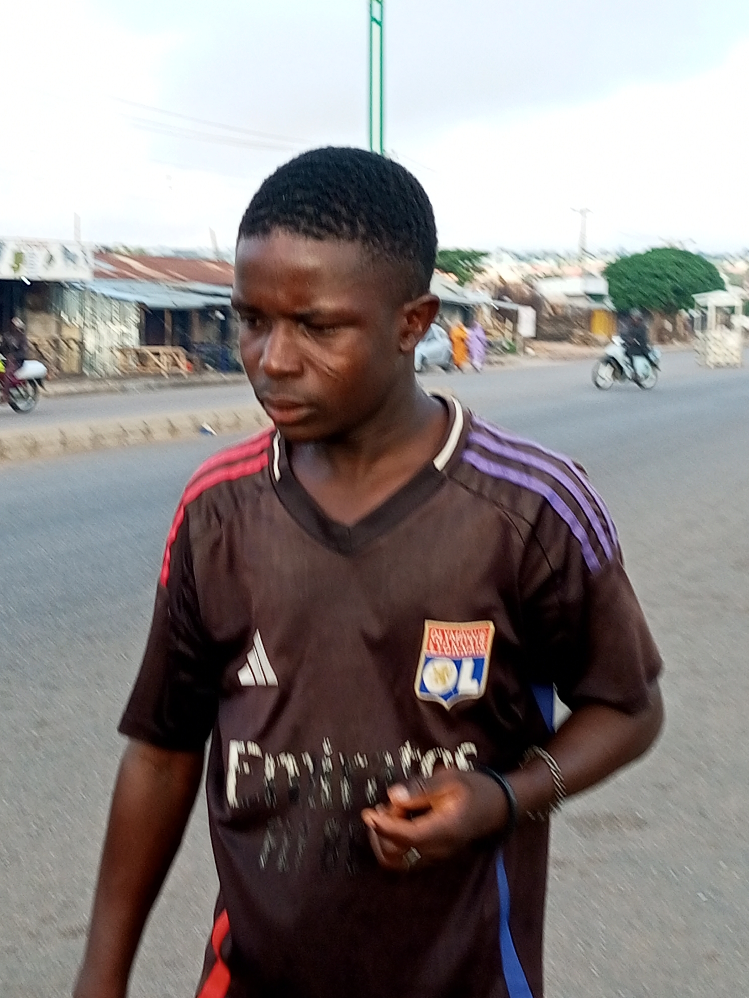 A medium shot captures a young Black male wearing a brown t-shirt with red and purple stripes on the sleeves and a crest on the chest, standing on a paved street in Gombe, Nigeria. He has a short, textured haircut and a scar on his left cheek. The t-shirt has a white Adidas logo on the left chest and the word "Emirates" printed across the front. He is wearing a beaded bracelet on his right wrist and a ring on his right index finger. The background shows a street with sparse vegetation and buildings on either side. To the left, a motorcycle with a rider is visible. To the right, a person on a motorcycle is also visible, with more distant buildings and trees in the background. The sky is overcast.