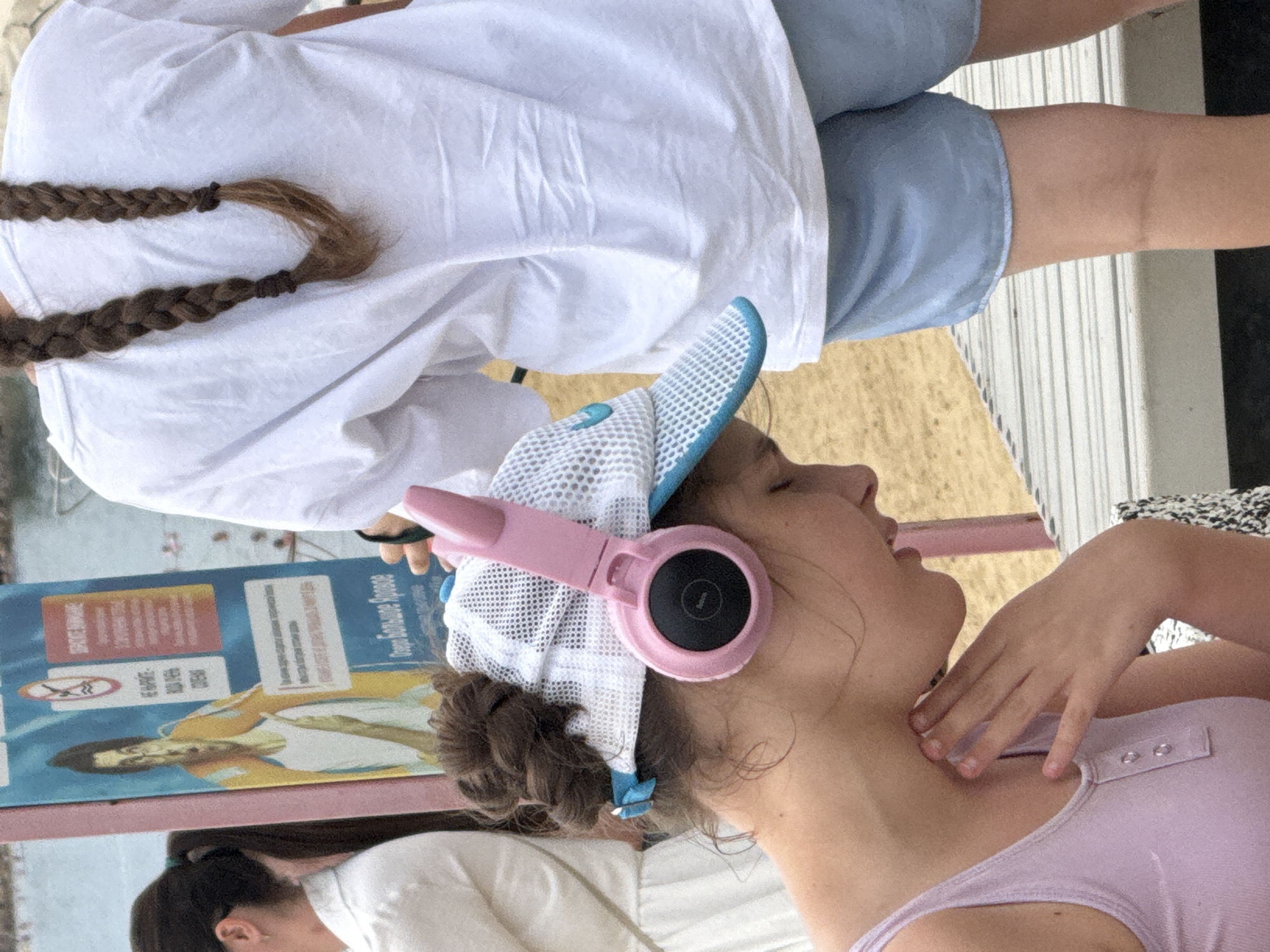 The image shows a close-up view of a young girl wearing a pink headset and a white and blue baseball cap. She is looking down and to the right, with her hand resting on her neck. She is wearing a light purple tank top. In the background, another person is visible wearing a white t-shirt and light blue shorts. They are standing in front of a colorful sign that appears to be a public notice or advertisement. The sign has an illustration of a person in an orange life jacket and some text in Russian. The environment appears to be outdoors, with sandy ground and a wooden platform visible. The overall impression is that of a casual outing in a public area, possibly a beach or park. The sign in the background mentions "озеро Большое Яровое," which translates to "Lake Bolshoye Yarovoye." This indicates the location is likely near or on the shore of Lake Bolshoye Yarovoye in Yarovoye, Russia.