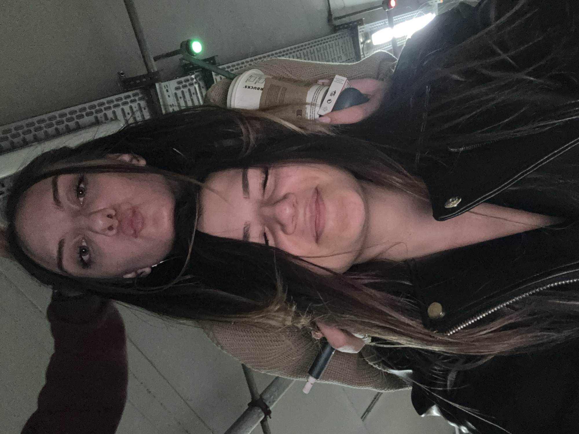 Two young women are posing closely for a selfie. The woman on the left has a pout expression, while the woman on the right is smiling with her eyes closed. The woman on the right is wearing a black leather jacket. In the background, there is a Starbucks coffee cup, an unidentified object, and a metal structure. The location appears to be indoors, possibly a parking garage, due to the overhead lighting and metallic elements. The precise location in Sofia, Bulgaria cannot be determined from the given image.