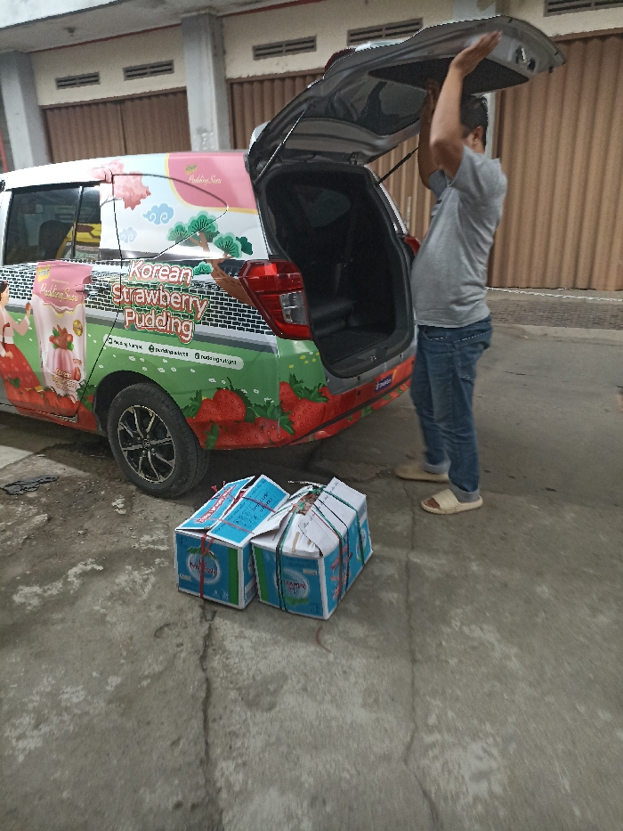 A man in a gray shirt and blue jeans is standing next to a white car with the back hatch open. The car is covered in colorful graphics depicting a strawberry pudding product with the words "Korean Strawberry Pudding" prominently displayed on the side. The man is holding onto the edge of the open hatch and appears to be about to close it. There are two boxes, possibly containing the product, lying on the gray concrete pavement in front of the car. The image is likely taken in Jatinangor, Indonesia, as the location is identified as coordinates -6.934, 107.775. The surrounding area is residential, with buildings, trees, and a paved street visible in the background.