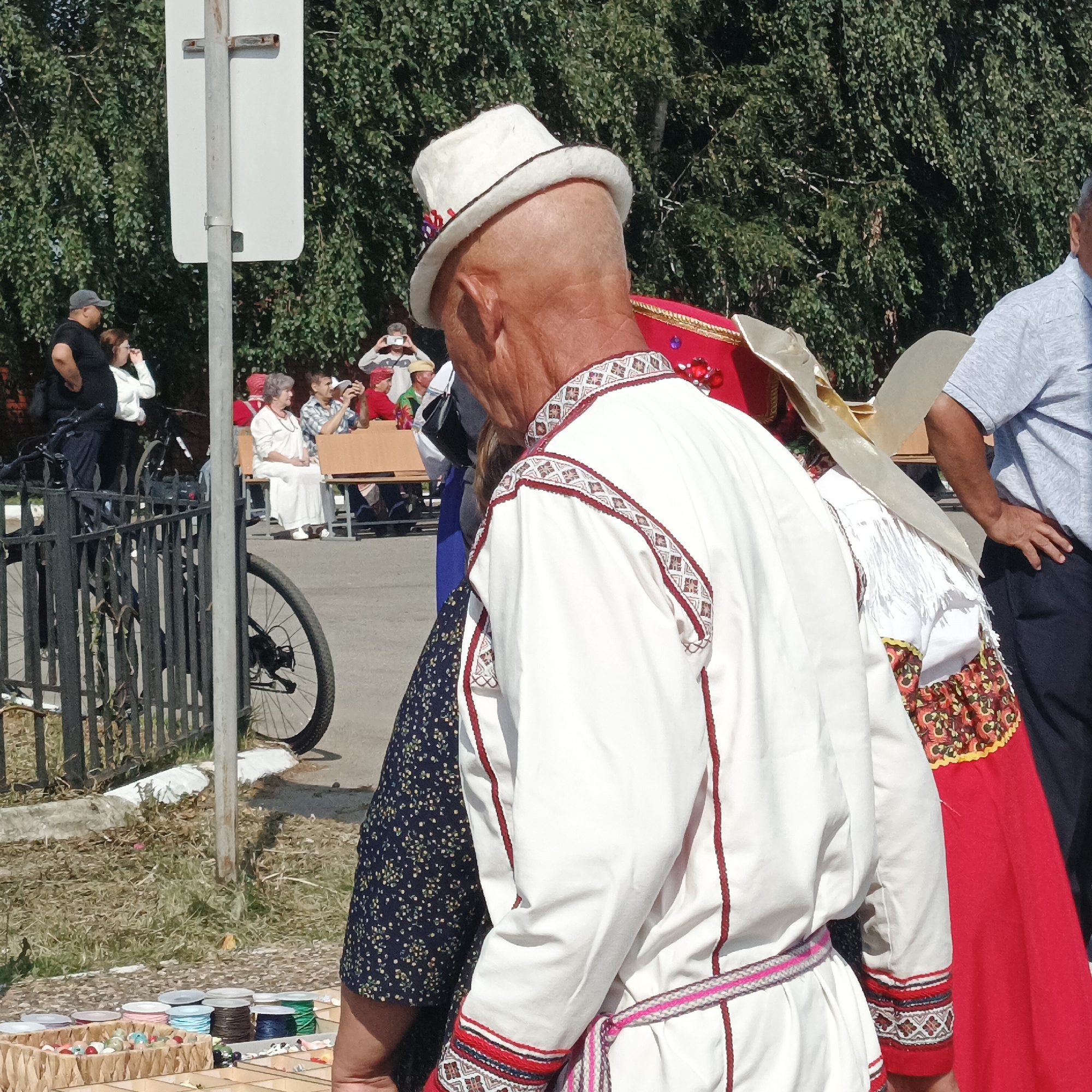 In Nikolo-Beryozovka, Russia, an outdoor scene unfolds with a focus on individuals in traditional attire. A man, viewed from the back, wears a white hat and a white, long-sleeved garment adorned with intricate red and white geometric embroidery along the collar and cuffs, and a braided pink and white belt. To his left, a woman in a dark blue, floral-patterned dress stands partially visible. Behind them, other people are present. A group sits on a wooden bench, some holding cameras, suggesting an event or performance. The environment includes lush green trees, a metal fence, a bicycle, and a display of craft items, including spools of thread and decorative beads, laid out on a surface in the foreground. A white utility pole stands prominently near the fence.