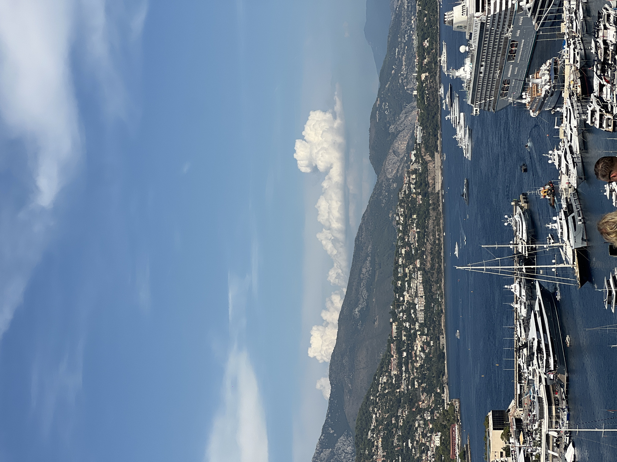 This is an elevated view of the Port Hercules in Monaco, taken on a clear day with blue skies and scattered clouds. The harbor is filled with numerous luxury yachts and a large cruise ship. The city of Monaco sprawls across the hills in the background, with buildings visible cascading down towards the water. The foreground shows several rows of moored boats, including a prominent dark-hulled mega yacht. In the distance, the mountainous coastline of France is visible. The water is a deep blue, reflecting the sky. Some people are visible in the lower right corner of the image, looking out at the harbor.