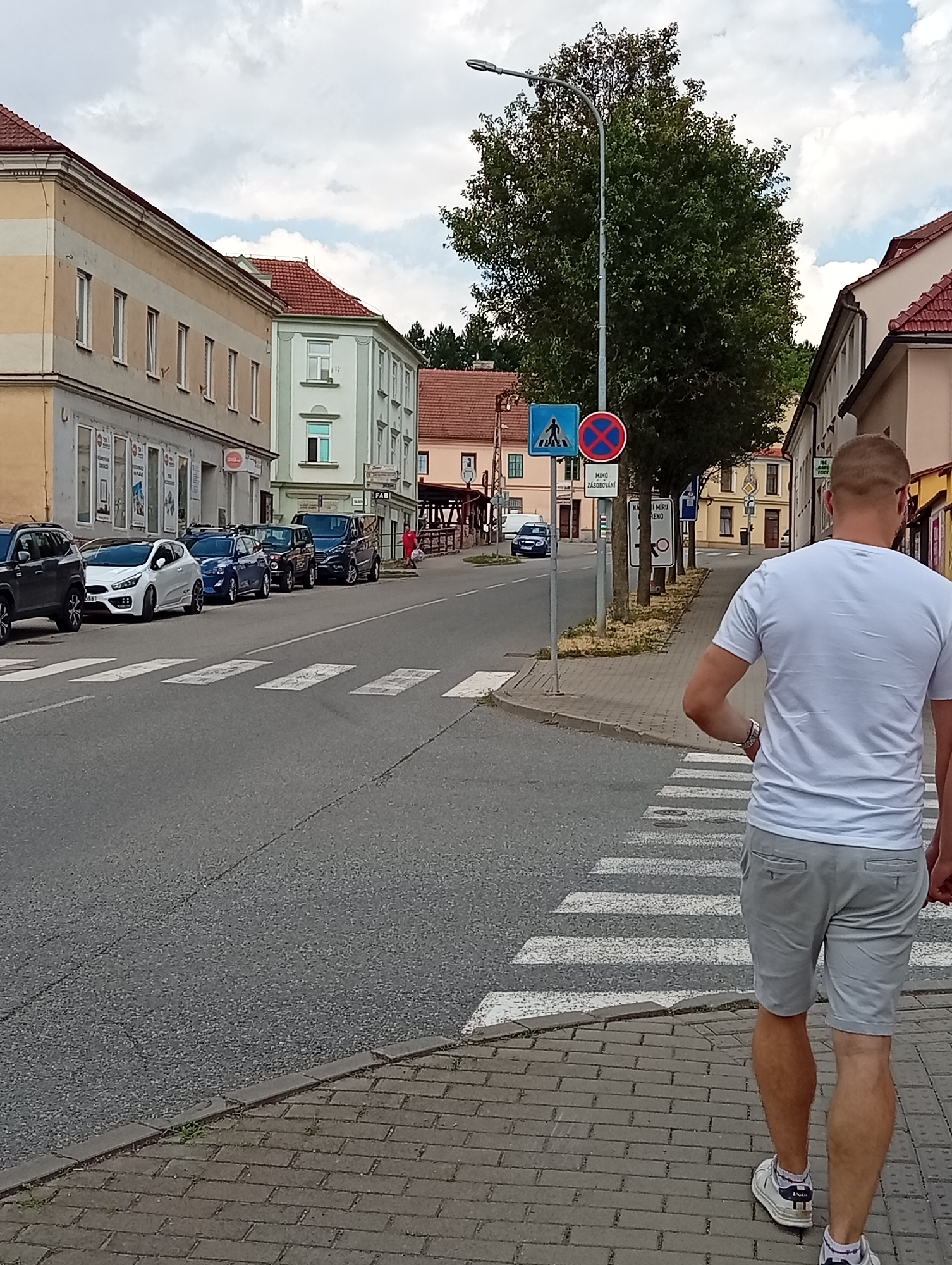 A person, seen from behind, walks on a brick sidewalk next to a street in Tišnov, Czechia. The person is wearing a white t-shirt and light grey shorts, with white sneakers and socks. They are walking away from the viewer and towards a pedestrian crossing. The street has several cars parked along the side and a crosswalk marked with white stripes. Buildings line the street, with a mix of architectural styles, including older structures with red tile roofs and more modern facades. Trees are visible, including a large one with dense foliage near a lamppost. Traffic signs are posted on the lamppost, including a pedestrian crossing sign and a circular sign with a red X, indicating "no parking" or a similar restriction. The sky is partly cloudy, suggesting daytime with moderate weather. The overall scene depicts a typical street in a European town.