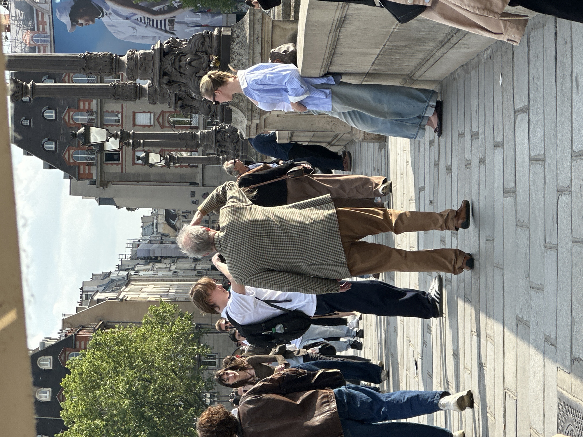 The image depicts a group of people standing on a stone bridge in Paris, France. The group is composed of various individuals, including a person wearing a tweed jacket, another wearing a blue button-down shirt, and others in casual attire. They are oriented towards the left side of the image, and some appear to be looking at something off-camera.
The bridge features ornate metal lamp posts and stone construction. Beyond the bridge, Parisian buildings with distinct architectural details, including arched windows and rooftops, can be seen. A large advertisement featuring a person is displayed prominently behind the buildings. A tree with green foliage is also visible, adding a natural element to the urban environment.