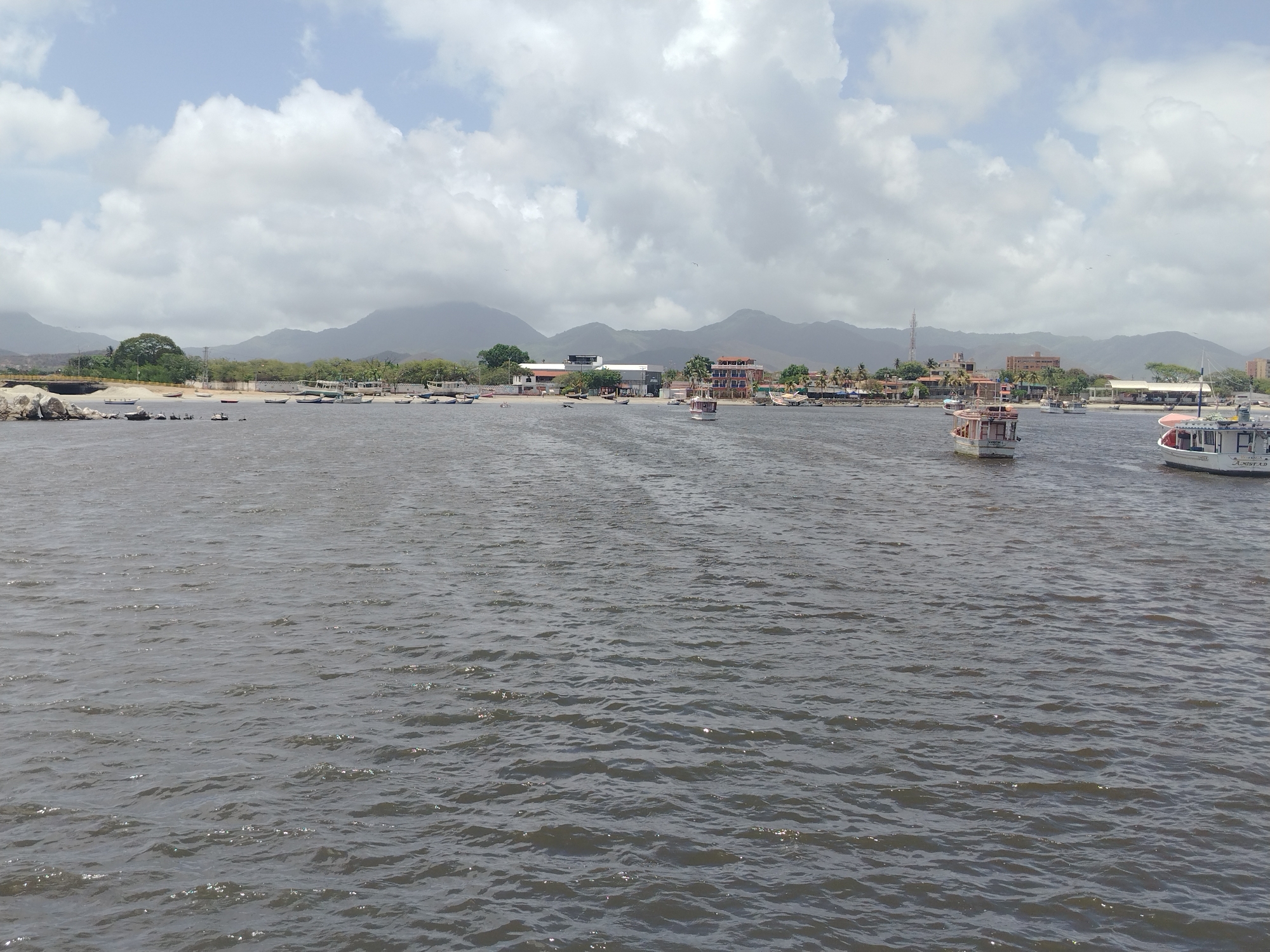 This media file depicts a scenic view of Juan Griego, Venezuela, a coastal town. The scene is dominated by a large body of dark, choppy water in the foreground, with a shoreline visible in the mid-ground. Along the shore, a collection of boats of various sizes are moored or beached. These boats are a mix of small fishing vessels and larger, more modern-looking boats.
In the background, the town of Juan Griego is spread out along the coast, characterized by a cluster of buildings with red and white roofs, and some taller structures that could be hotels or apartments. Behind the town, majestic mountains rise, their peaks softened by a hazy, overcast sky. The sky itself is filled with a mix of white, puffy clouds and lighter grey clouds, suggesting a dynamic weather pattern.
The time of day appears to be daytime, likely late morning or early afternoon, given the brightness of the sky. The weather seems partly cloudy and possibly warm, typical of a coastal climate. There are no people prominently featured in the image, but the presence of numerous boats suggests a fishing or maritime community. The overall mood is one of tranquility and natural beauty, with a touch of human activity evident in the harbor. There is some visible text on one of the boats on the right side of the frame, but it is too blurry to be legible.