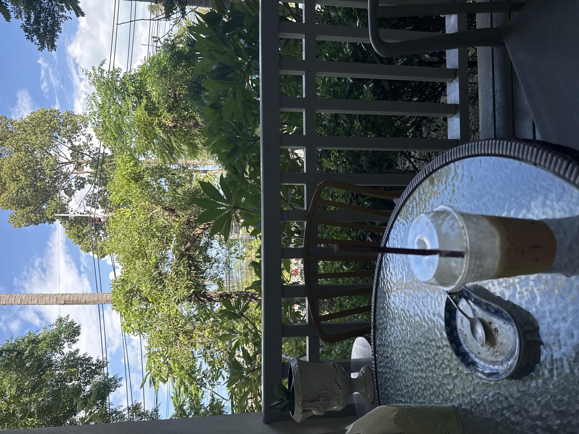 This is a close-up shot of a table on an outdoor balcony overlooking lush greenery in Ko Samui District, Thailand. A clear plastic cup with a straw, filled with a light brown beverage and ice, sits on the glass table. Next to it is a small, blue and white patterned dish with a spoon, showing remnants of food. A white napkin dispenser is visible on the left edge of the table. In the background, a white railing separates the balcony from a dense garden with various trees and bushes. The sky above is blue with scattered white clouds.