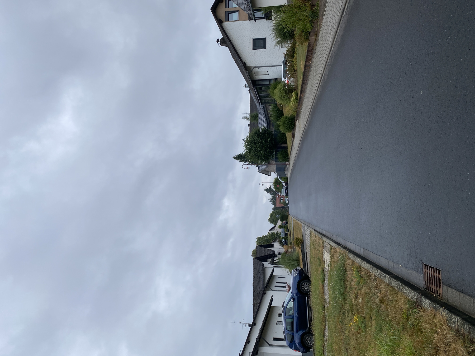 The image depicts a residential street in the city of Flammersfeld, Germany, under an overcast sky. On the left side of the street, a dark blue SUV is parked on a patch of dry grass adjacent to a row of white houses with dark-tiled roofs. The houses have minimalist facades with small windows. A grey asphalt road dominates the center of the image, curving slightly towards the distance. On the right side of the road, more residential houses are visible, characterized by white textured walls and dark roofs, interspersed with green bushes and trees. A few other vehicles are sparsely parked along the street. The ground alongside the road features a grey paved sidewalk and patches of grass. A metal storm drain grate is visible on the left side of the road near the foreground. The overall scene is quiet and suburban.