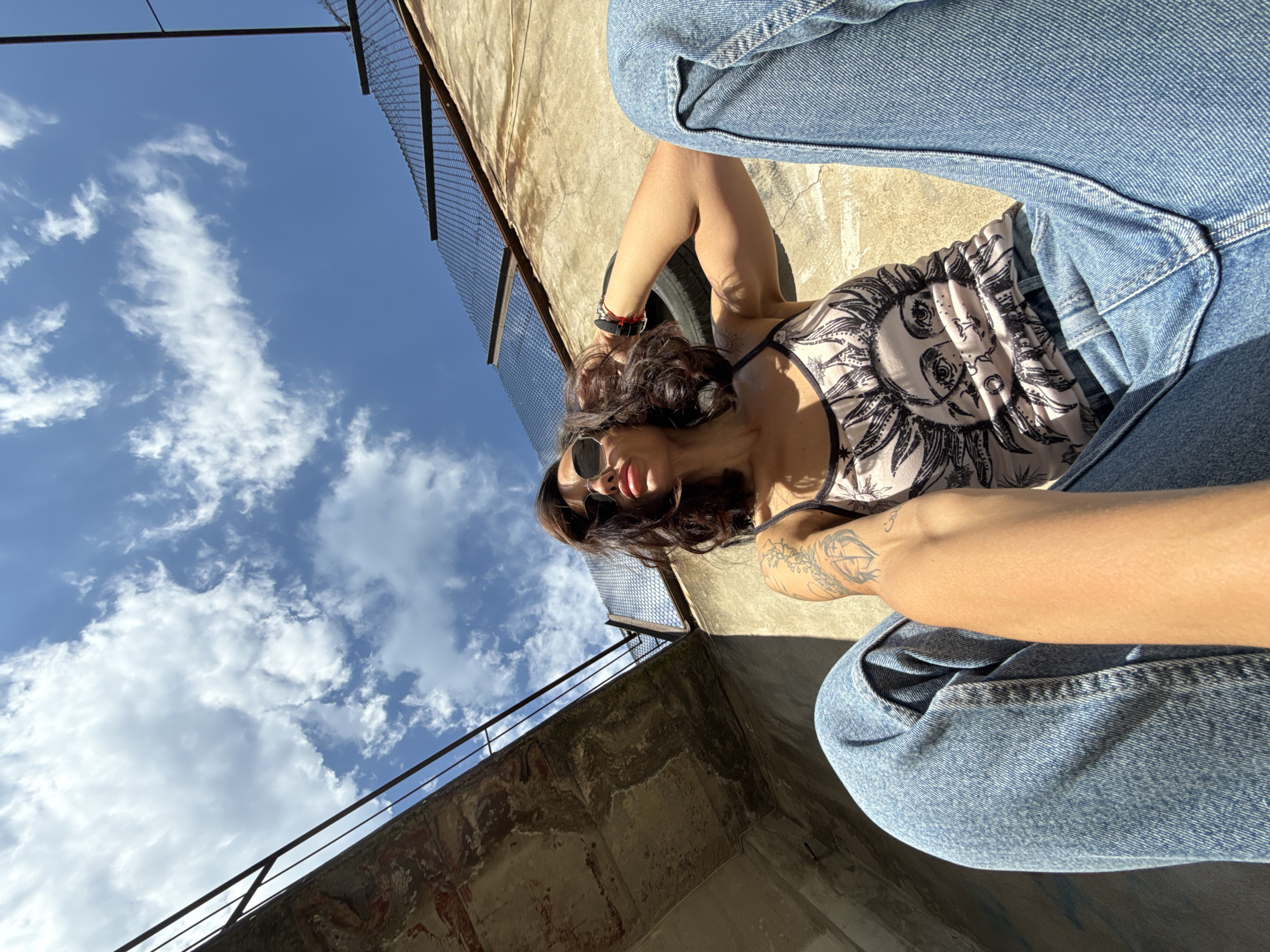 A woman is sitting outdoors in Pleven, Bulgaria. She is wearing sunglasses, a sleeveless shirt with a sun design, and light blue jeans. The sky is blue with some clouds. There's a partially visible weathered building with a metal railing/fence to the left and a taller building with a wire fence at the top to the right. The woman appears to be leaning back against something unseen.