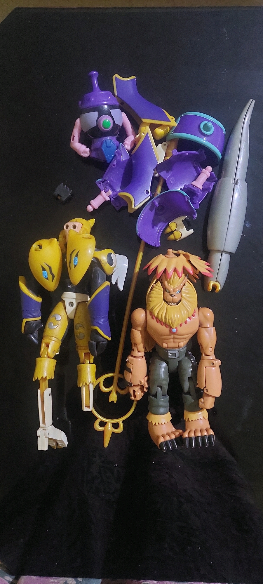The image displays a collection of action figures arranged on a dark, textured surface. In the upper portion of the image, several disassembled purple and yellow plastic toy pieces are scattered, including a small character with a purple head and pink arms, and larger components that appear to be armor or decorative elements. To the right of these pieces is a long, silver, pointed object, possibly a weapon or accessory.
Below these disassembled parts, two complete action figures stand. The figure on the left is predominantly yellow and purple, with a stylized, somewhat insectoid or robotic appearance. It has large yellow shoulder pads and yellow limbs with purple accents. The figure on the right is a bipedal character with a lion-like head, a muscular orange torso, dark green pants, and large, clawed feet. It is holding a gold staff with a circular element.
The overall arrangement suggests a collection of toys, possibly for display or play, laid out on a dark background that could be a table or floor. The lighting is somewhat uneven, with shadows cast by the figures and objects. There is no indication of any specific location in Lumajang, Indonesia.
