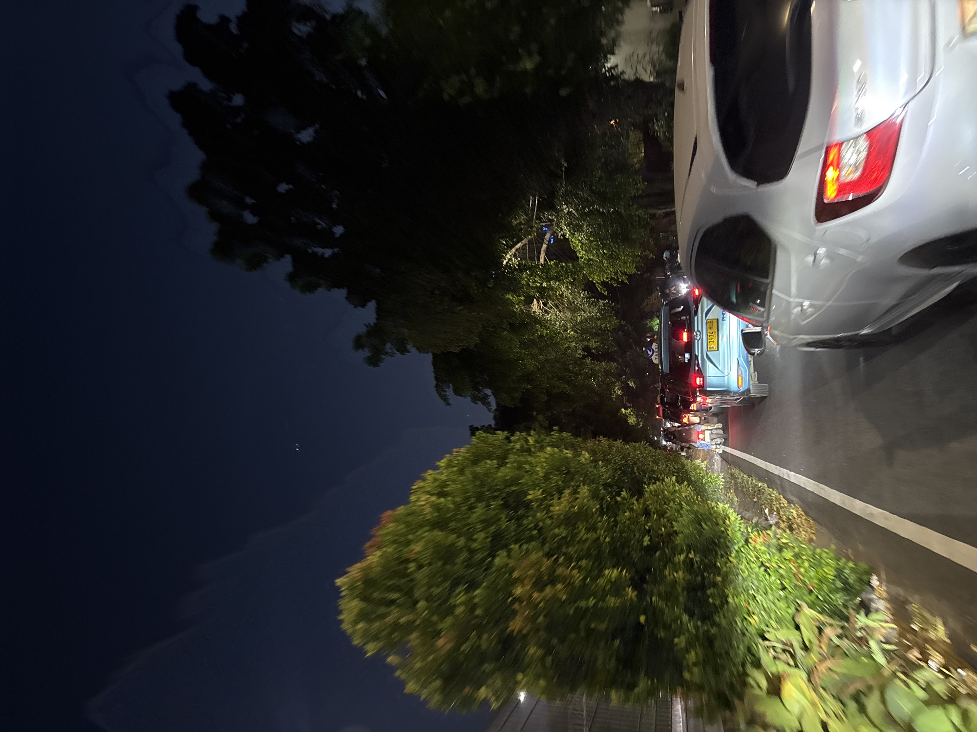 The image depicts a traffic scene at night, likely on a street in West Jakarta, Indonesia. The perspective is from within a vehicle, looking forward. A white car, possibly a hatchback, is in the foreground, its rear taillight visible. Ahead, a light blue vehicle with a license plate starting with "S-1914" is also in traffic. Several motorcycles are interspersed within the congestion, their headlights and taillights illuminated. Dense green trees line the right side of the road, with a lighter, more manicured hedge on the left. The sky above is dark blue, with some blurry cloud formations and faint lights that could be stars or distant city lights. The overall impression is of slow-moving traffic on a tree-lined road at night.