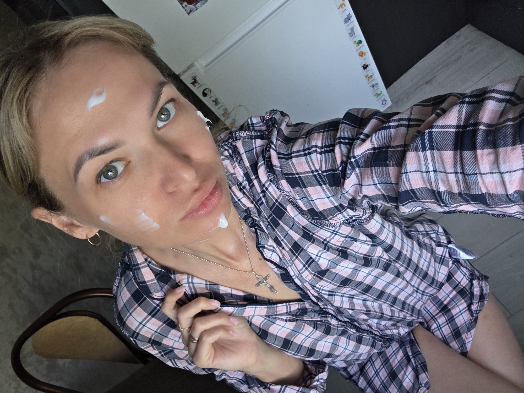 A woman with blonde hair is taking a selfie indoors. She has dots of white cream on her forehead, cheek, and chin. She is wearing a pink and black plaid shirt, a gold hoop earring, and a necklace with a cross pendant. An earbud is visible in her ear. A white refrigerator with colorful animal stickers is in the background. The setting appears to be a home in the city of Chaikovsky, Russia.