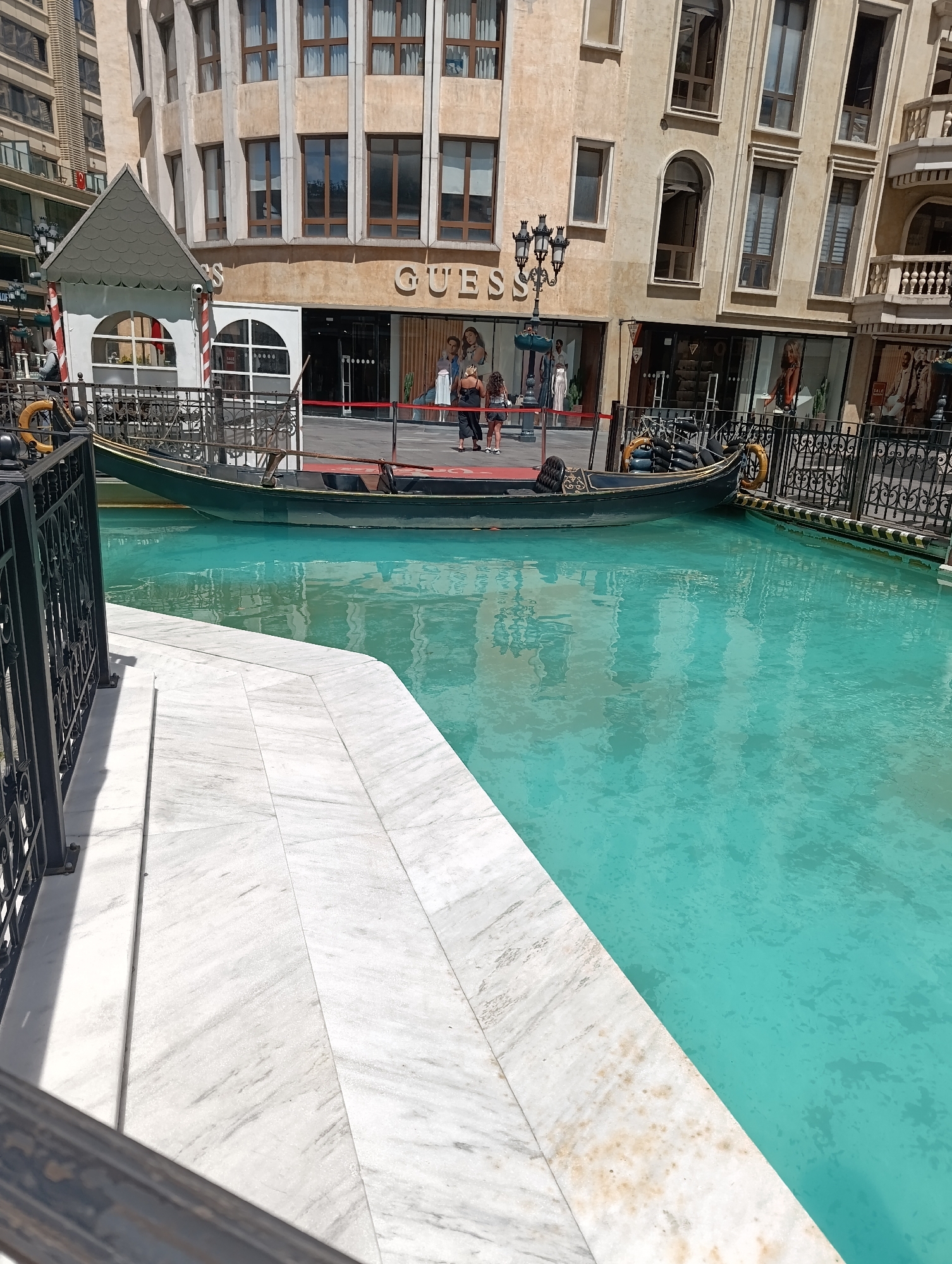 The image depicts a scene at the Venezia Mega Outlet mall in Gaziosmanpaşa, Turkey, which is designed to resemble Venice. A dark-colored gondola, characteristic of Venetian canals, floats on vibrant turquoise water. The water is calm, with reflections of the surrounding buildings visible on its surface. The buildings themselves have an architectural style that mimics Venetian structures, with beige-colored facades, arched windows, and decorative balconies. A prominent "GUESS" store is visible on one of the buildings, with mannequins displayed in the storefront windows. A lamppost with multiple lights stands near the entrance of the store. In the foreground, a white marble pathway with a textured pattern runs alongside the water, bordered by a black metal railing with ornate scrollwork. A small, white, gabled structure, resembling a ticket booth or small kiosk, is also present near the water's edge. Two individuals are partially visible near the "GUESS" store, appearing to be walking. The overall scene aims to evoke the ambiance of Venice within an urban shopping environment.