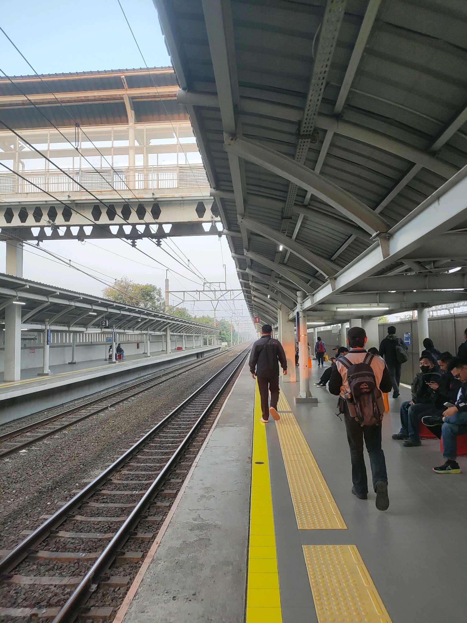 The image depicts a train station platform in Telagamurni, Indonesia. The platform is covered by a modern, grey, corrugated metal roof supported by a structure of white steel beams. Parallel train tracks run alongside the platform, which has a grey concrete surface with yellow tactile paving near the edge. Several people are visible on the platform, some walking, some seated on benches. In the foreground, two men are walking away from the camera. The man on the left wears a dark jacket and trousers, while the man on the right wears a dark jacket, dark trousers, and a backpack. In the background, other individuals are waiting or walking, and some are seated on benches. The environment appears overcast, with a clear sky visible above the station roof. The station signage is partially visible, including a sign with the number "2" indicating a platform. Overhead electrical wires for the trains are visible. The overall scene is one of a functional, contemporary urban transportation hub.