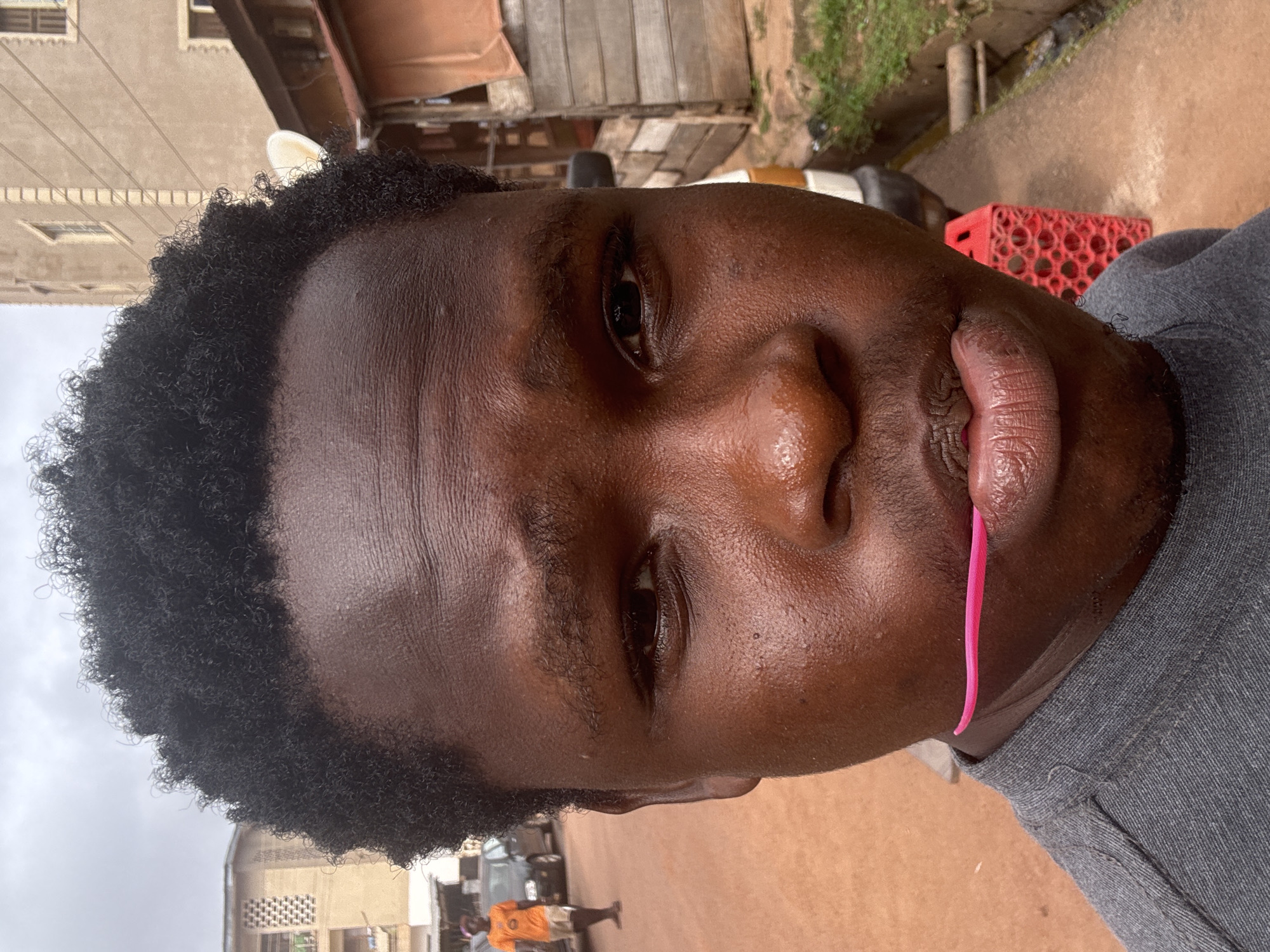 The image is a close-up portrait of a young Black man with curly black hair, taken outdoors in what appears to be a street scene in Enugu, Nigeria. The man is wearing a grey t-shirt and has a bright pink object, possibly a toothpick or a decorative hair accessory, held between his lips. His skin is dark and slightly shiny, and he has a serious expression.
In the background, there is a building with several windows and balconies, characteristic of urban architecture in Nigeria. To the right, there's a wooden structure with a corrugated iron roof, and a red plastic object with a honeycomb-like design. A paved walkway with some vegetation and a drainage channel is visible. Further in the background, there are parked cars and another person wearing an orange shirt and shorts, walking away from the camera. The ground is reddish-brown, suggesting the presence of laterite soil, common in the region. The sky is overcast, indicating a cloudy day. The overall setting suggests an everyday urban environment in Enugu.