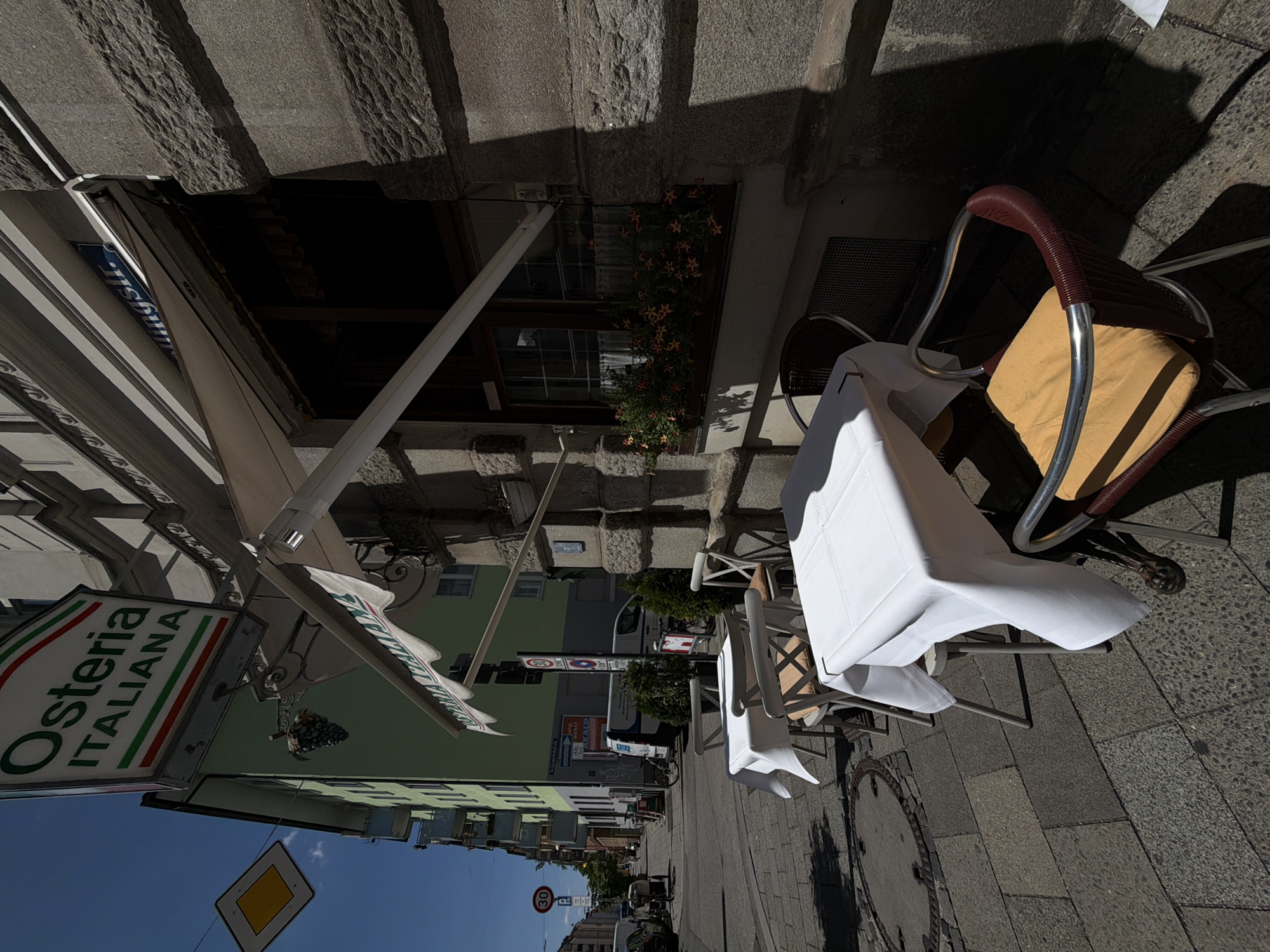 The image depicts an outdoor seating area of "Osteria Italiana" located on Lindwurmstraße in Munich, Germany. Two tables are set with white tablecloths, accompanied by chairs. One table is partially obscured by shadow, while the other is in full view, with a yellow cushioned chair positioned at it. A building with a light green facade and a stone exterior stands behind the tables. A sign above the building reads "Osteria Italiana" in green and white lettering. Street signs and traffic signs are visible in the background. The scene is illuminated by bright sunlight, casting distinct shadows on the cobblestone pavement.