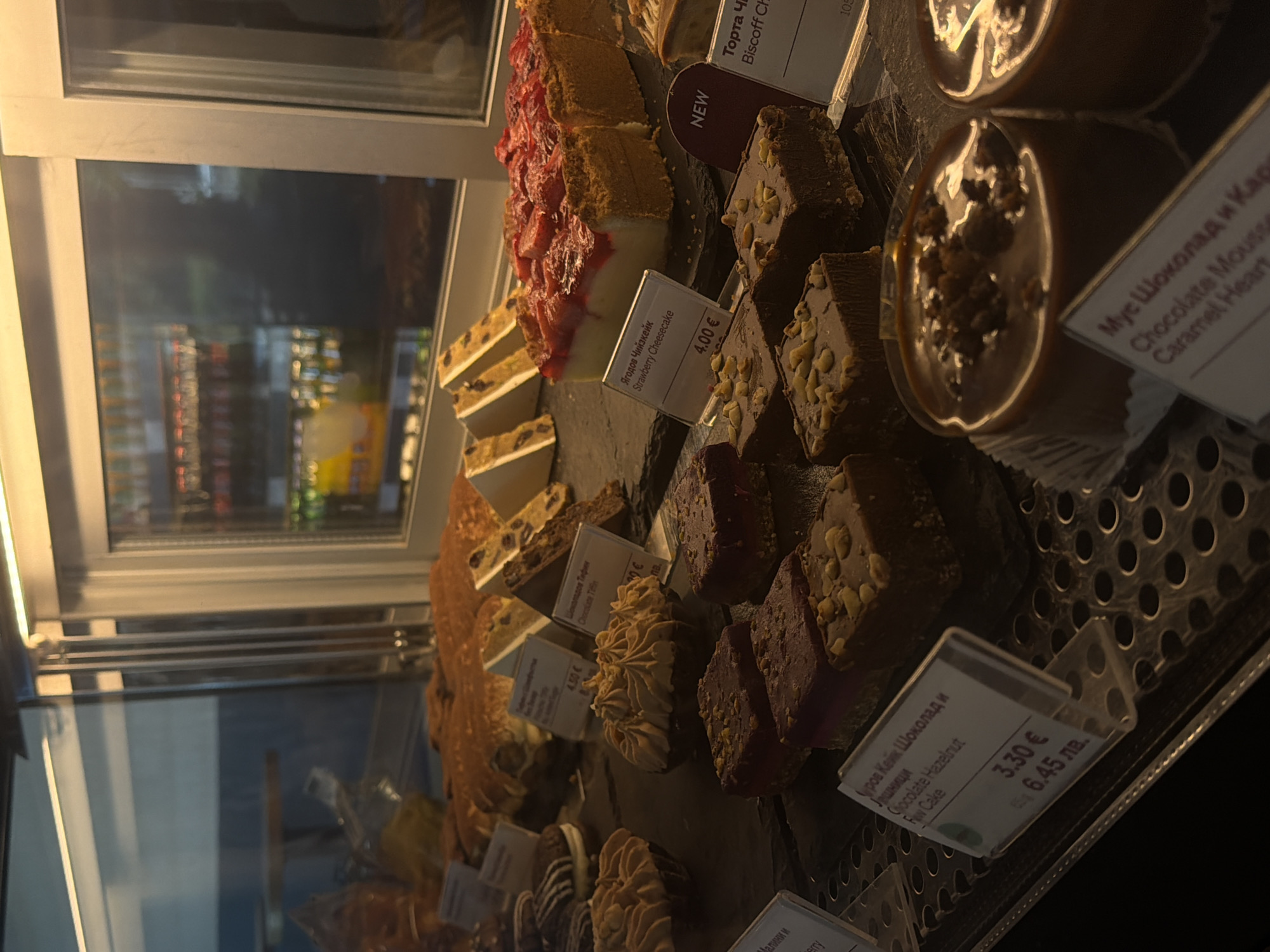 In a bakery display case, various desserts are arranged on tiered shelves. The case features a bright overhead light that illuminates the baked goods. In the foreground, a row of small chocolate cakes with a glossy caramel topping and crumbles are visible. To their left, several square chocolate brownies topped with nuts are displayed on a dark surface. Further back, a strawberry cheesecake with a crumb crust and a layer of creamy filling is cut into portions. Behind it, slices of fruitcake and other pastries are visible. The display case has labels with product names and prices in a foreign language and Euros. The background shows the interior of the bakery, including shelves stocked with bottles and other products. The overall lighting is warm and inviting, highlighting the appetizing appearance of the desserts.
