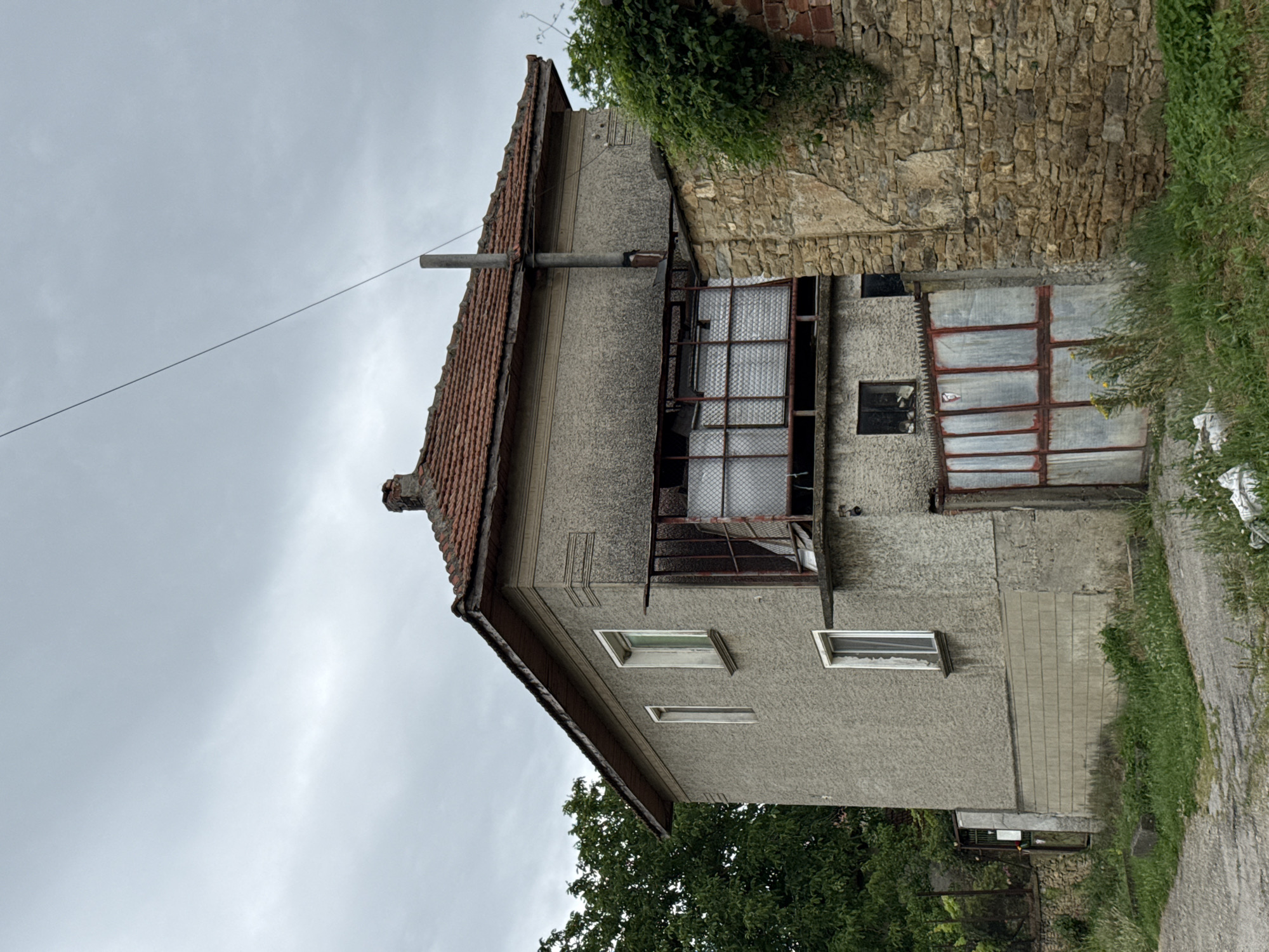 The image depicts a two-story, weathered concrete house with a red tiled roof, situated in the city of Slavyani, Bulgaria. The house has several windows, some of which appear to be boarded up or covered with mesh. A metal balcony structure is visible on the second floor, also covered with mesh. Adjacent to the house is a section of an old stone wall, partially overgrown with vegetation. A metal gate with a red frame and reflective panels is positioned in front of the lower level of the house. The surrounding area is characterized by overgrown grass and a dirt or gravel pathway. The sky is overcast with gray clouds. There are no people visible in the image, and no discernible actions are taking place. The scene appears to be an exterior view of a residential property in a rural or semi-rural setting.