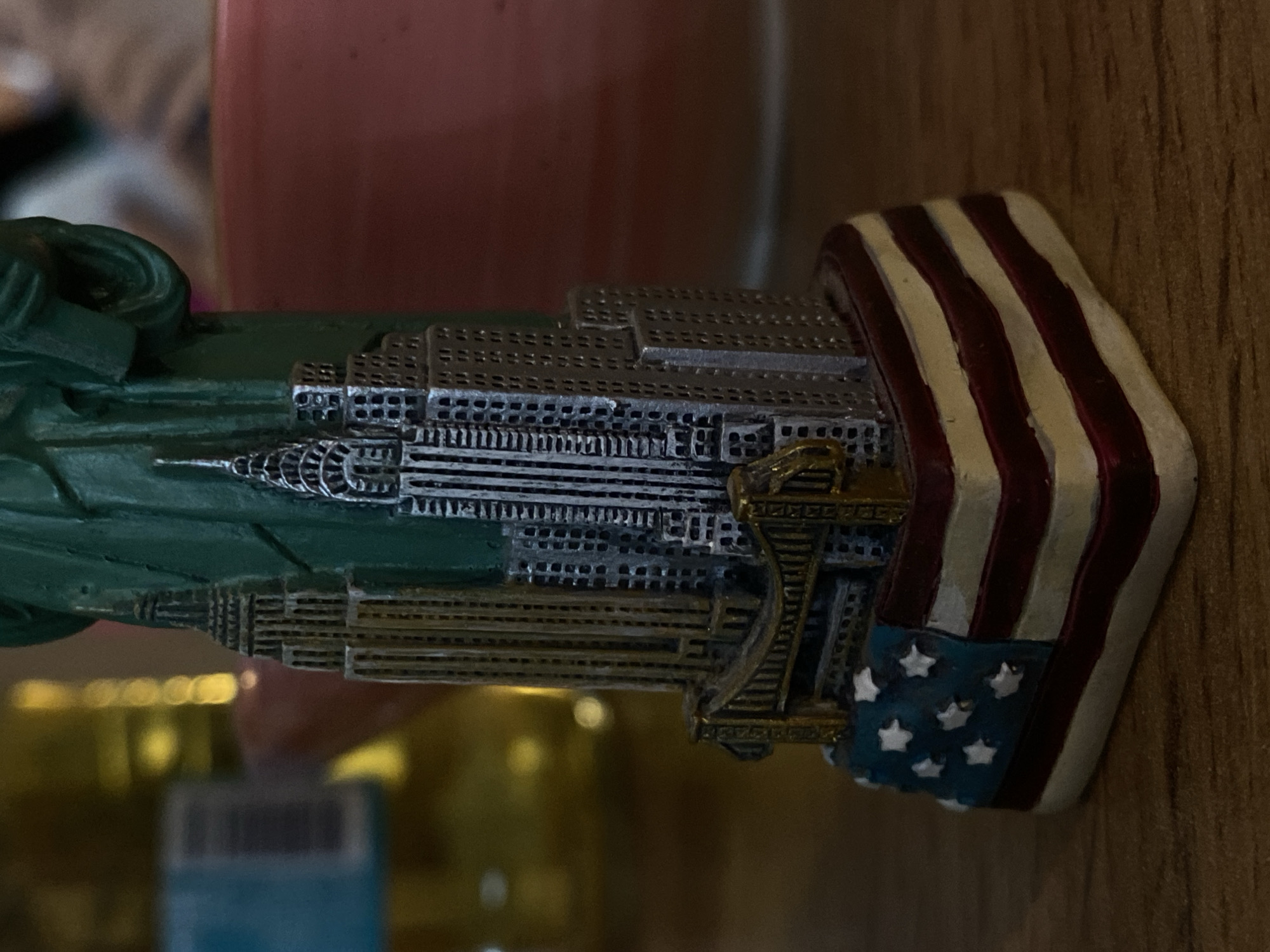 The image shows a souvenir depicting the Statue of Liberty, the New York City skyline, and the American flag. The souvenir is positioned horizontally against a wooden surface. The Statue of Liberty is green, and the buildings of the skyline are predominantly silver. The American flag component displays red and white stripes with a blue field and white stars. The location is not in Votkinsk, Russia.