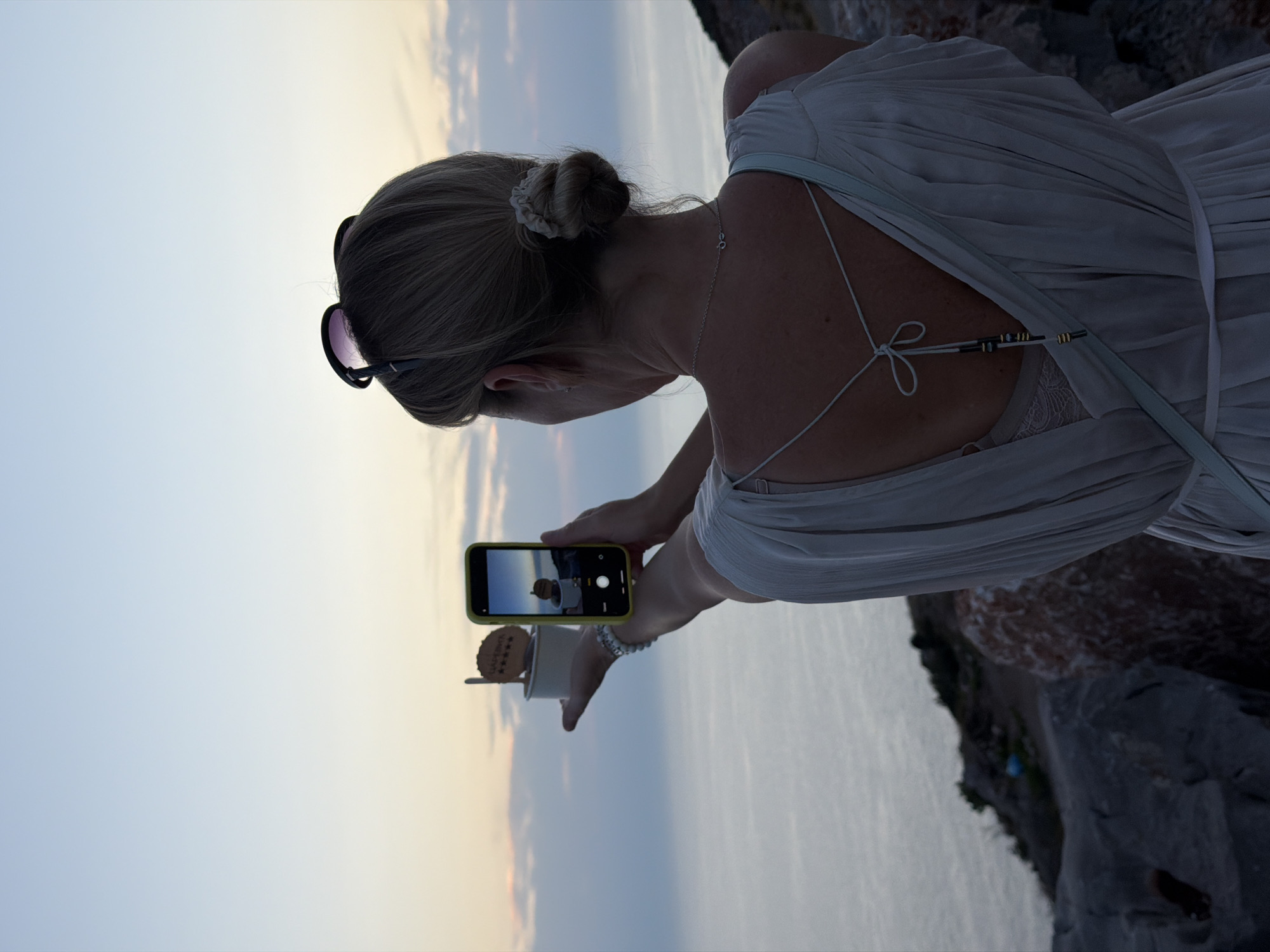 A woman with blonde hair pulled back in a bun is seen from behind, holding a smartphone in her right hand to take a picture of a small ice cream in a white cup with a wooden stick and a decorative wooden disk. She is wearing a light-colored, flowing dress and sunglasses perched on her head. The background shows a serene sea and a soft, twilight sky. The scene is set on a rocky cliffside overlooking the water, suggesting a location like Kovačko Polje, Montenegro.