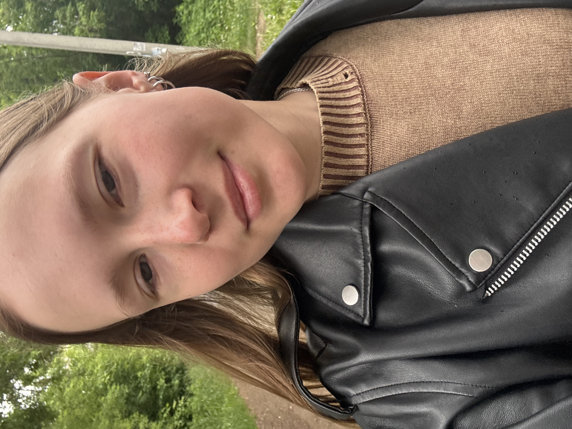 A young individual with long, light brown hair, a light complexion, and visible freckles on the nose and cheeks, is depicted in a close-up, chest-up shot. The person is looking slightly downward and to their left, with a subtle smile and closed lips. The left ear is visible, adorned with two small silver hoop earrings. The individual wears a black faux leather biker-style jacket featuring silver snap buttons and a prominent silver zipper. Beneath the jacket, a tan or light brown knitted sweater with a ribbed collar displaying horizontal darker brown stripes is visible, along with a thin silver chain necklace. The background consists of dense green foliage, consistent with trees and bushes, indicating an outdoor setting. A dirt or gravel path is partially visible to the left, and a light-colored utility pole stands in the right background. The scene occurs in natural daylight, in the city of Chaikovsky, Russia.