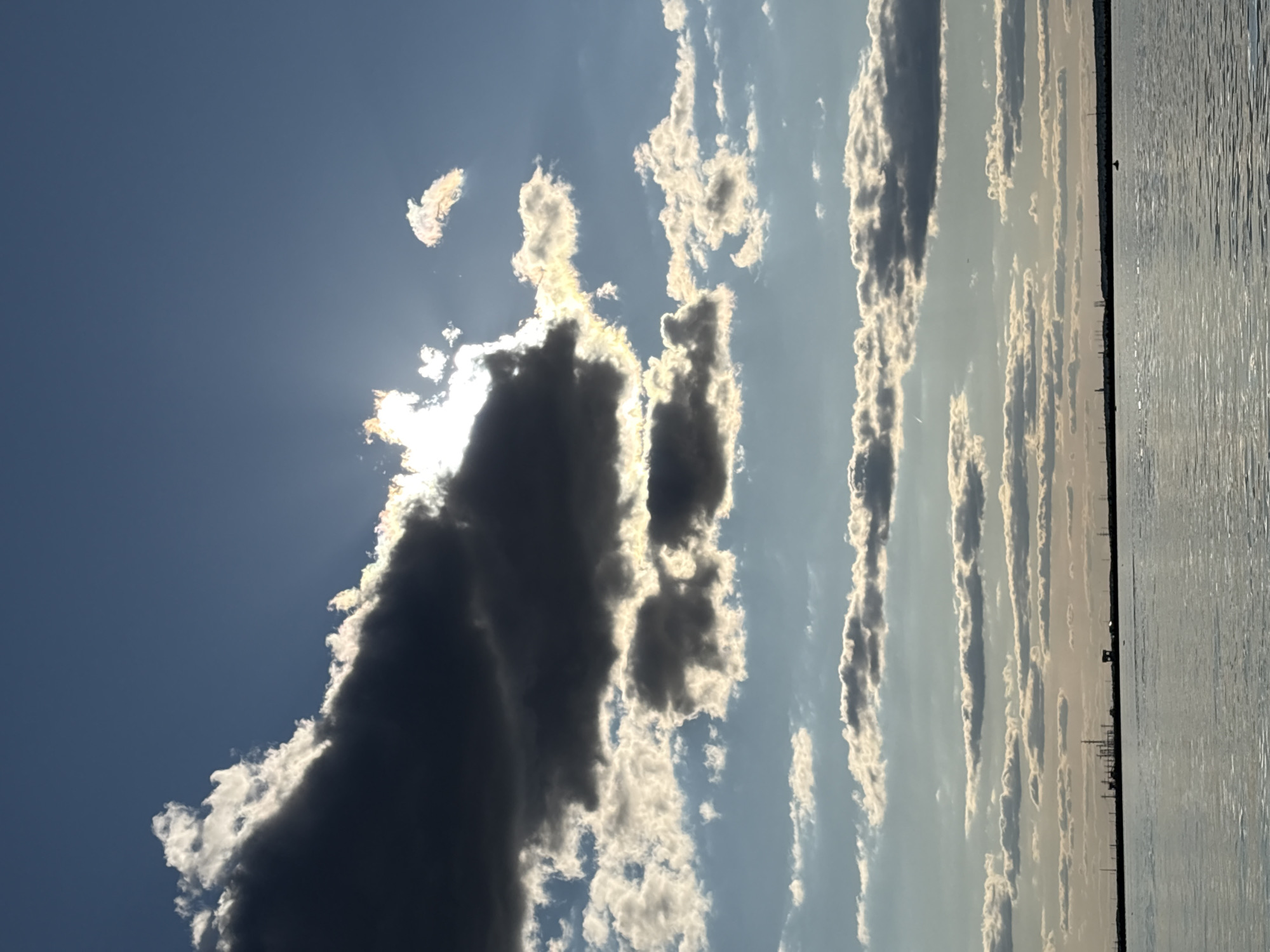 The image depicts a wide-angle view of a body of water, likely a river or bay, under a dramatic sky. The water is calm with subtle ripples reflecting the light. The horizon is marked by a distant, flat shoreline, dotted with industrial structures and silhouetted objects that suggest a port or industrial area. The sky is a dominant feature, filled with a variety of clouds. Large, dark storm clouds are positioned in the upper left, partially obscuring the sun, which creates bright rays of light and highlights the edges of the clouds with a golden glow. Smaller, more scattered white and gray clouds are present throughout the sky, some appearing thinner and more wispy, while others are more substantial. The overall impression is one of a dynamic and somewhat moody sky.