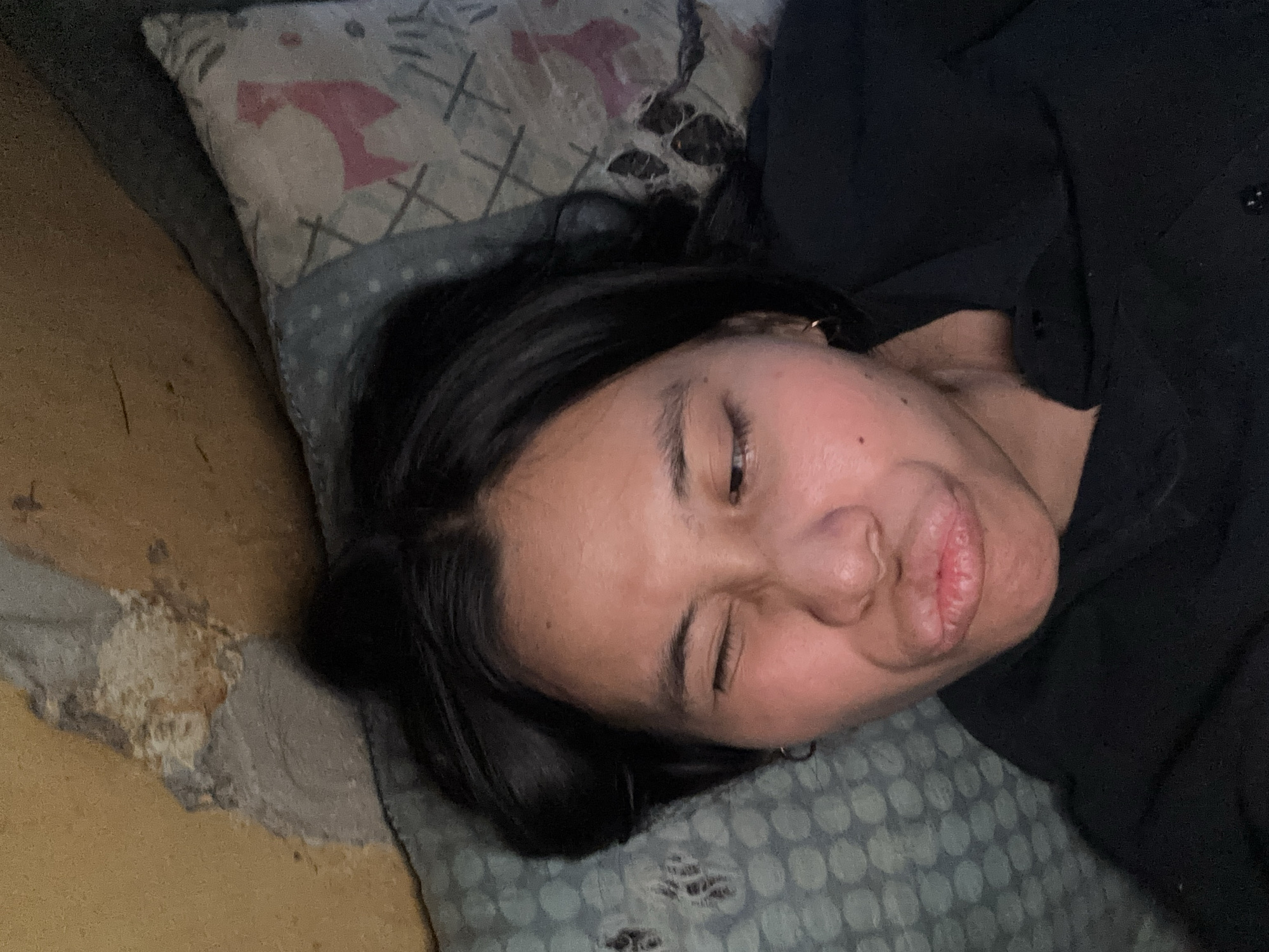 The image shows a close-up of a person's face as they lie down, with their mouth puckered and one eye winking. They are wearing a black shirt and have dark hair. The background appears to be a bed with patterned pillows and a textured wall or ceiling above. The scene is dimly lit, suggesting an indoor setting, possibly a bedroom. The details of the environment are not specific enough to pinpoint a location within East Jakarta, Indonesia.
