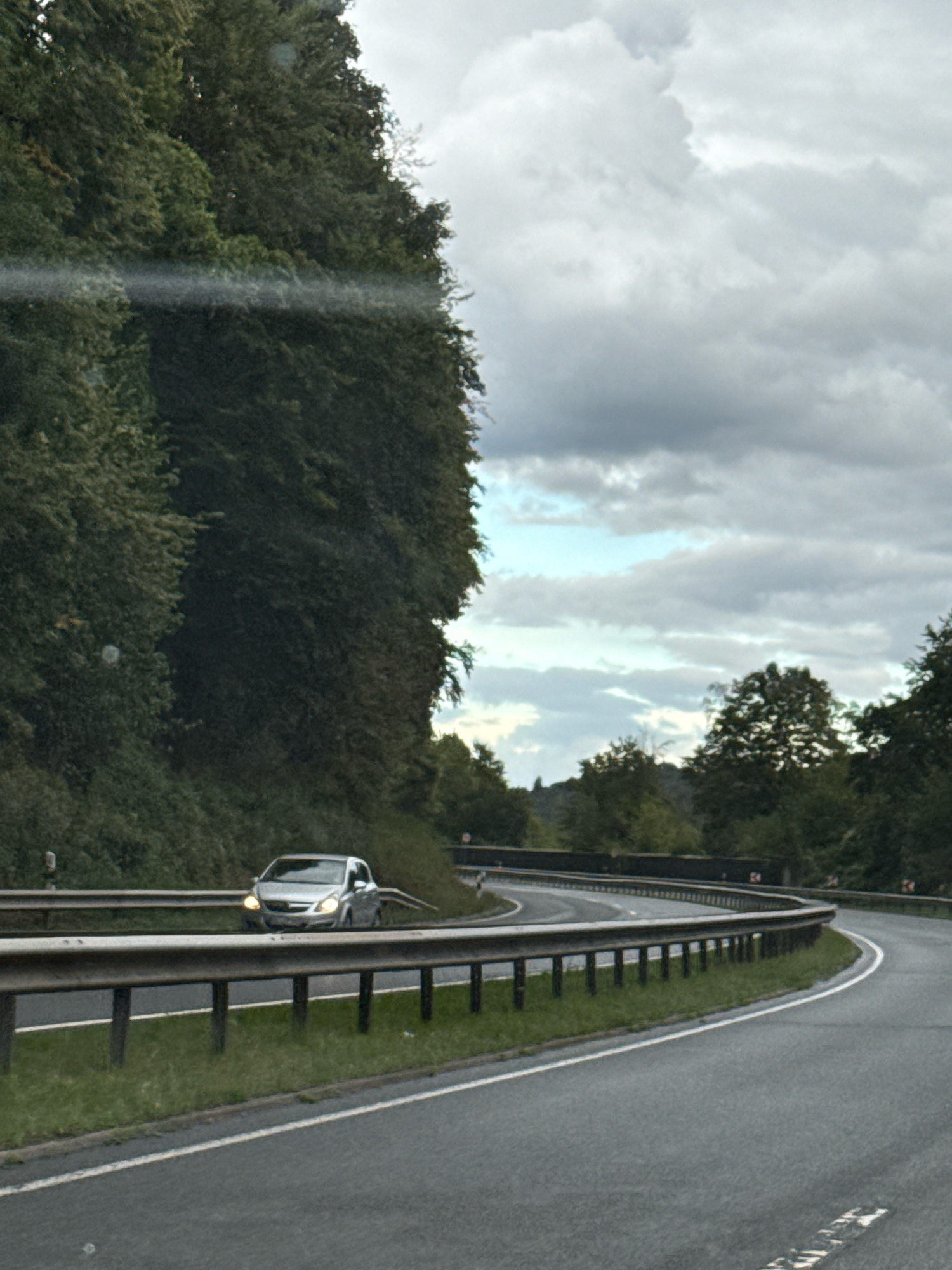 A silver car is driving on a winding road bordered by a metal guardrail and lush green trees. The sky is overcast with a mix of gray clouds and patches of blue. The road appears to be in a rural or semi-rural area, possibly a highway or a main road leading into or out of Herzberg, Germany, given the dense foliage and the style of the guardrail and road markings. There are no people visible in the frame. The lighting suggests it is daytime, but the clouds indicate potential for rain or a generally muted light. The car is positioned on the left side of the road, navigating a curve.