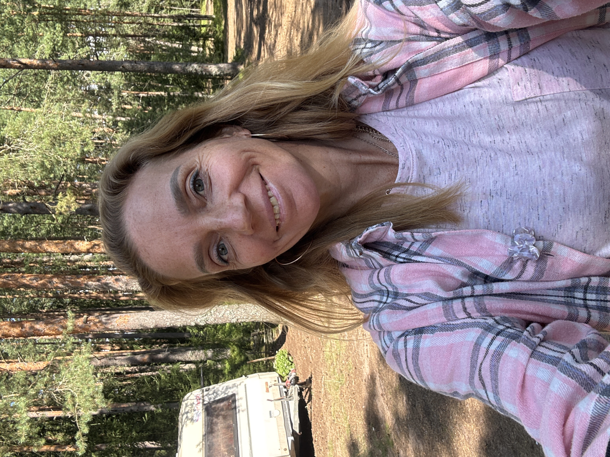 A woman is taking a selfie in a forest clearing. She has blonde hair and is smiling. She is wearing a pink and gray plaid shirt over a light pink t-shirt and several necklaces. She has hoop earrings. In the background, a white caravan is parked, and tall pine trees are visible, with a dirt path winding through them. The ground is covered in dry grass and pine needles. The image appears to have been taken during daylight hours.