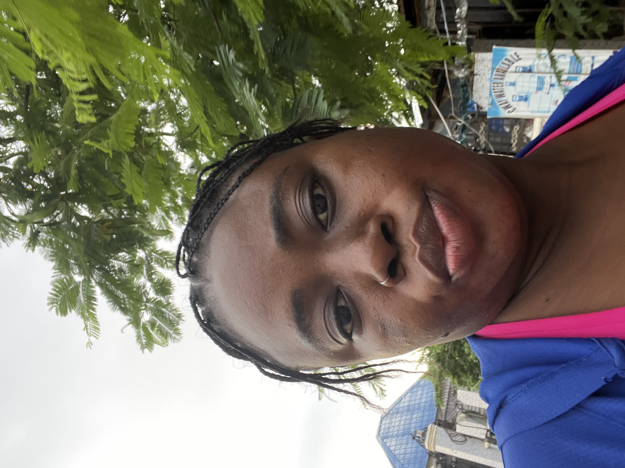 A close-up, selfie-style, outdoor shot shows a Black woman with braided hair and a nose ring. She is wearing a blue hooded jacket over a bright pink top. Her face is turned slightly to the right, and she has a playful expression.
In the background, lush green foliage from a tree hangs over the scene. To the left, a building with a distinctive blue, tiled roof is visible, along with an ornate gate. To the right, a sign for "GWAY WATER AVAILABLE" with images of water bottles is displayed. The overall environment suggests a moderately urban setting, likely in Abuja, Nigeria, possibly a residential or commercial area with outdoor spaces. The lighting indicates an overcast day.